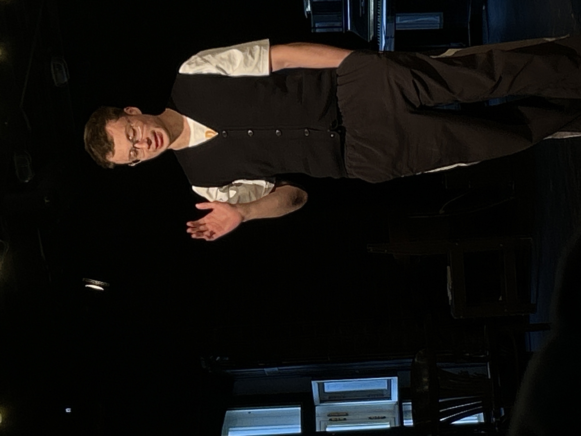 The image captures a young adult male performing on what appears to be a dimly lit stage or performance area. He stands upright, facing slightly towards the right side of the frame. He wears a white t-shirt, a dark, likely black, button-up vest over it, and dark trousers. A pair of clear-rimmed glasses is perched on his nose. His left hand is raised slightly with the palm open, and his mouth is ajar, suggesting he is speaking, singing, or mid-expression during a performance.
The background is predominantly dark, almost completely black, characteristic of a theatrical setting designed to focus attention on the performer. Faint hints of stage lighting or structural elements can be discerned in the upper left background. In the bottom left corner, two illuminated rectangular panels, possibly resembling windows or light-up stage props with horizontal details, offer a small contrast to the dark environment. The scene depicts an indoor event, likely a play, concert, or presentation, with the performer well-lit by unseen stage lights. There is no visible text in the image.