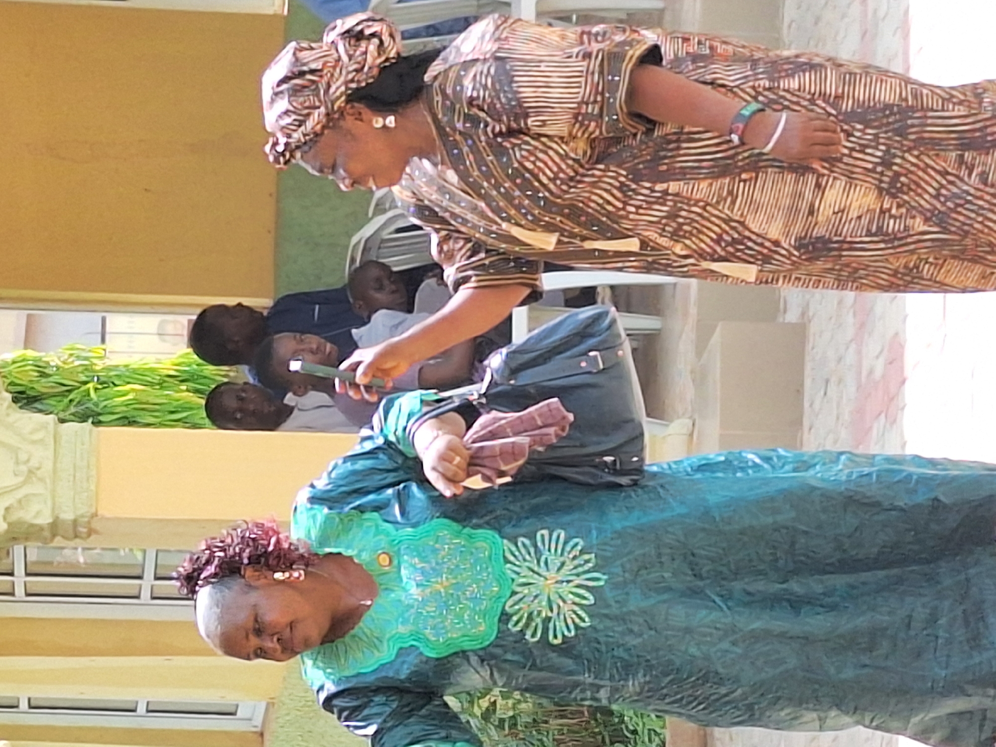 Two women stand in what appears to be an outdoor gathering. One woman wears a brown and beige patterned dress with a matching headwrap. She is looking down at a mobile phone being held by a younger person, possibly a child. Behind them, several other people are seated. The other woman, in the foreground, wears a green dress with floral embellishments. She is holding a cloth or handkerchief. The location is in Rido, Nigeria. The environment includes a building with windows and decorative elements, as well as some greenery.