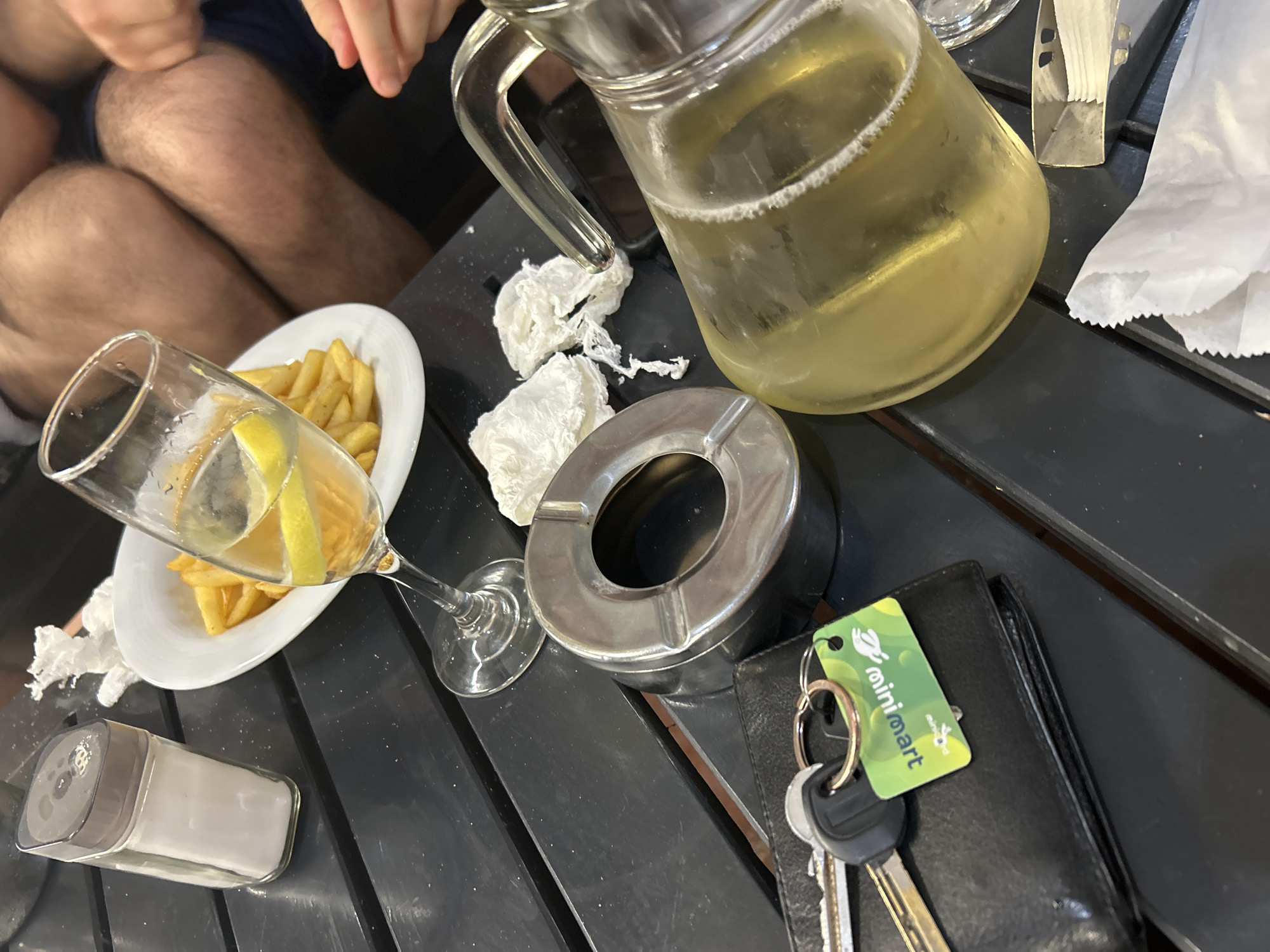 The image depicts an overhead view of a casual outdoor table setting in the city of Blagoevgrad, Bulgaria. The table is dark, with visible slats, suggesting a standard outdoor cafe or restaurant surface.
Prominently featured are multiple items for dining and leisure. Towards the upper left, a rectangular clear glass salt shaker with a grey cap stands next to a white, oval plate containing a portion of golden-brown french fries. Adjacent to the plate, a stemmed glass holds a light yellow beverage, garnished with two lemon slices.
In the central area of the table, a silver-colored metallic ashtray is positioned alongside several crumpled white paper napkins or tissues. To the right, a clear glass pitcher, filled with a pale yellow liquid exhibiting a frothy top, rests on the table. A portion of an unused white paper napkin is visible at the bottom right.
In the lower left, a black leather bi-fold wallet is placed on the table. Attached to a keyring on the wallet are two keys: a standard metal key and a black plastic car key/fob. A green rectangular keychain with a white rabbit logo and the text "minimart" (in English) and "אורורה" (in Hebrew) is also on the keyring.
In the background, the hairy knees and upper thighs of a person, consistent with a male wearing dark shorts, are partially visible, indicating someone seated opposite the viewpoint. A hand is also partially visible on the right side of the frame. The overall scene suggests a social gathering at an establishment where food and drinks are served, and smoking may be permitted.