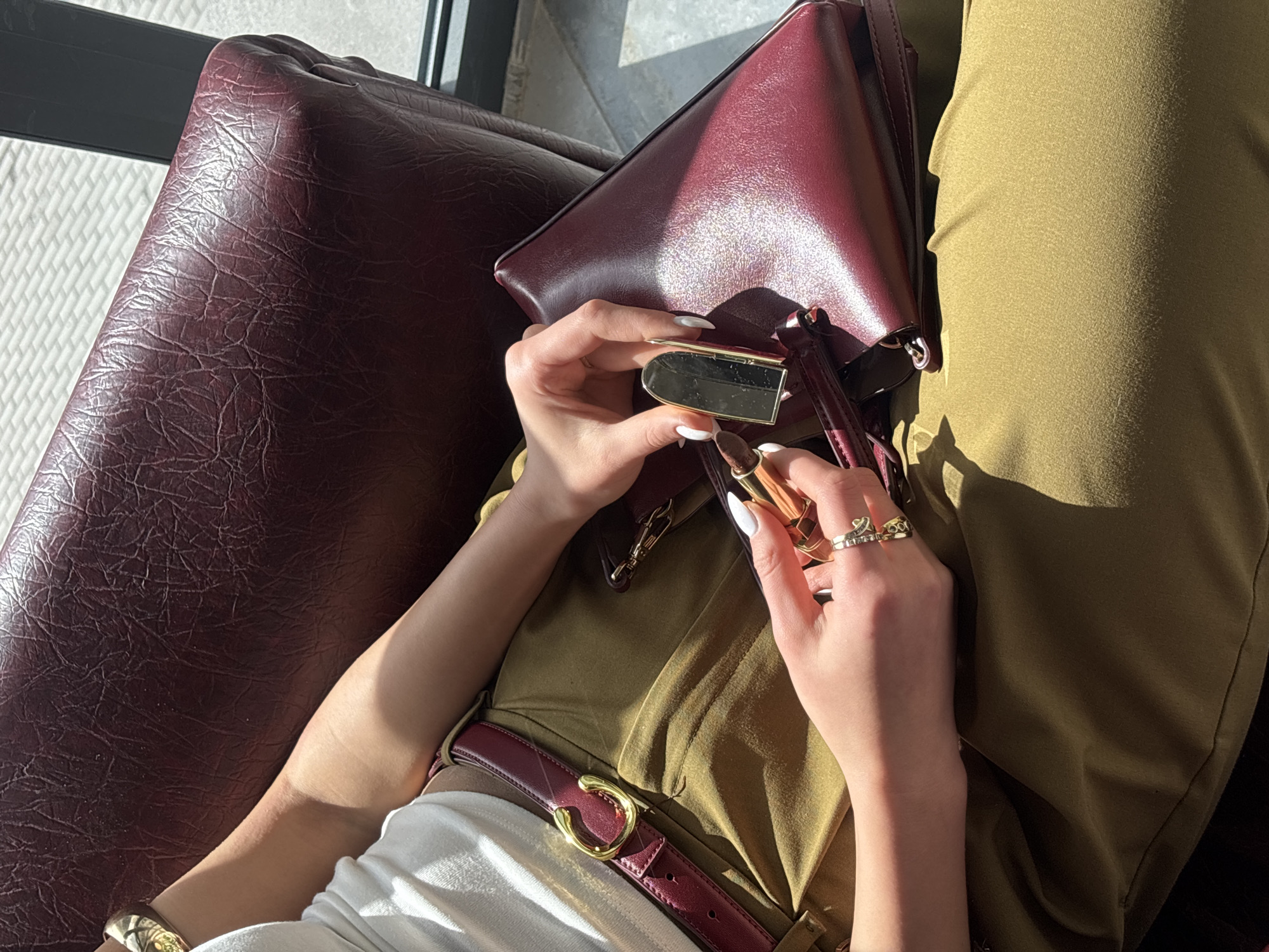 A woman sits on a burgundy leather chair in a sunlit area. She is holding a gold lipstick container in her left hand, its lipstick component exposed. She is opening the mirrored case with her right hand. She is wearing gold rings and a bracelet. A matching burgundy leather purse sits next to her on the chair. She is wearing olive-green trousers with a matching burgundy leather belt. The location is potentially a cafe or indoor space in Yerevan, Armenia, though there is no definitive city-specific evidence.