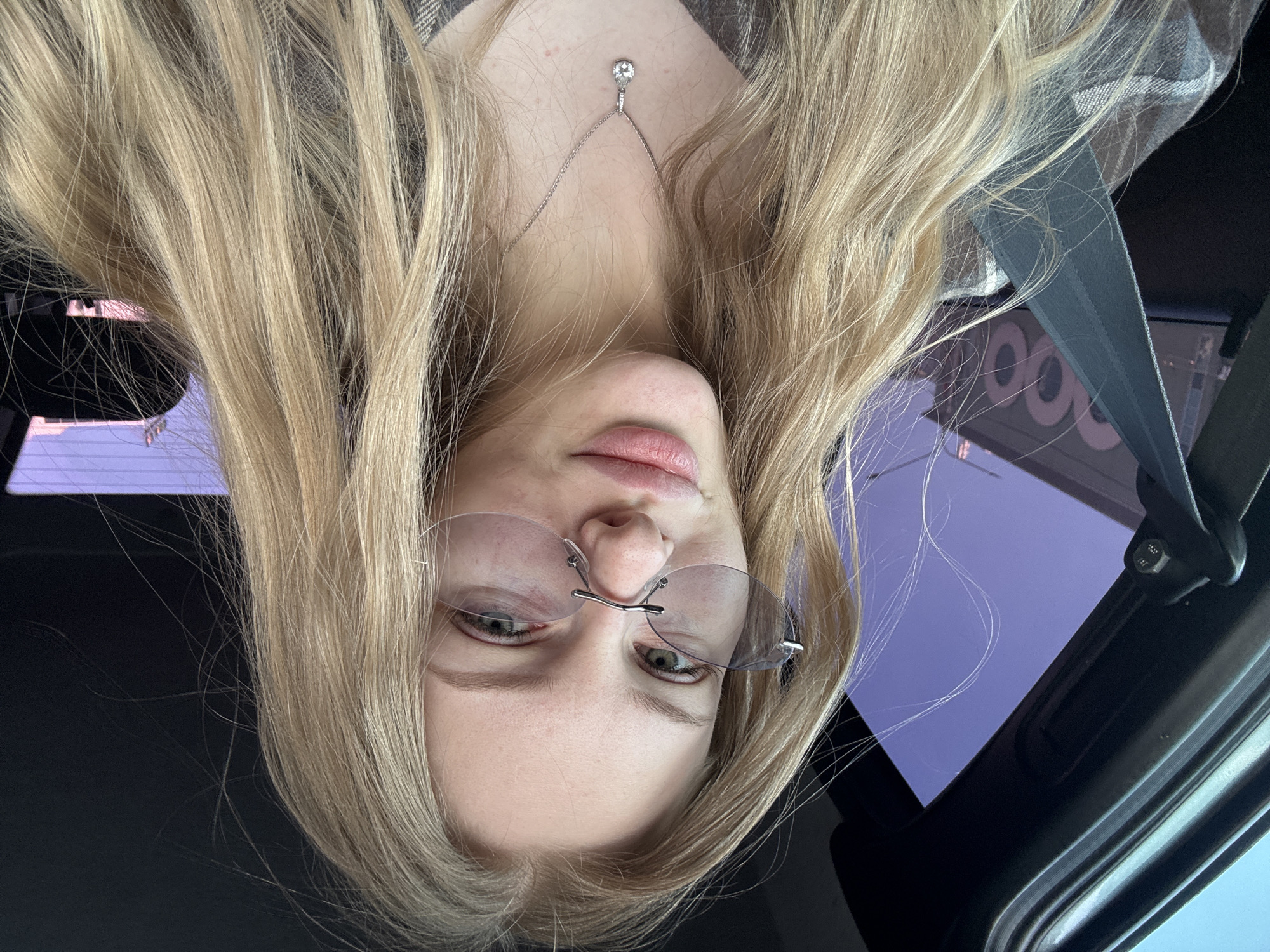 A woman with blonde hair and glasses is in the front seat of a car, wearing a seatbelt and a necklace with a pendant. Through the car window, a building is visible with the number "600" on it. The location is Perm, Russia.