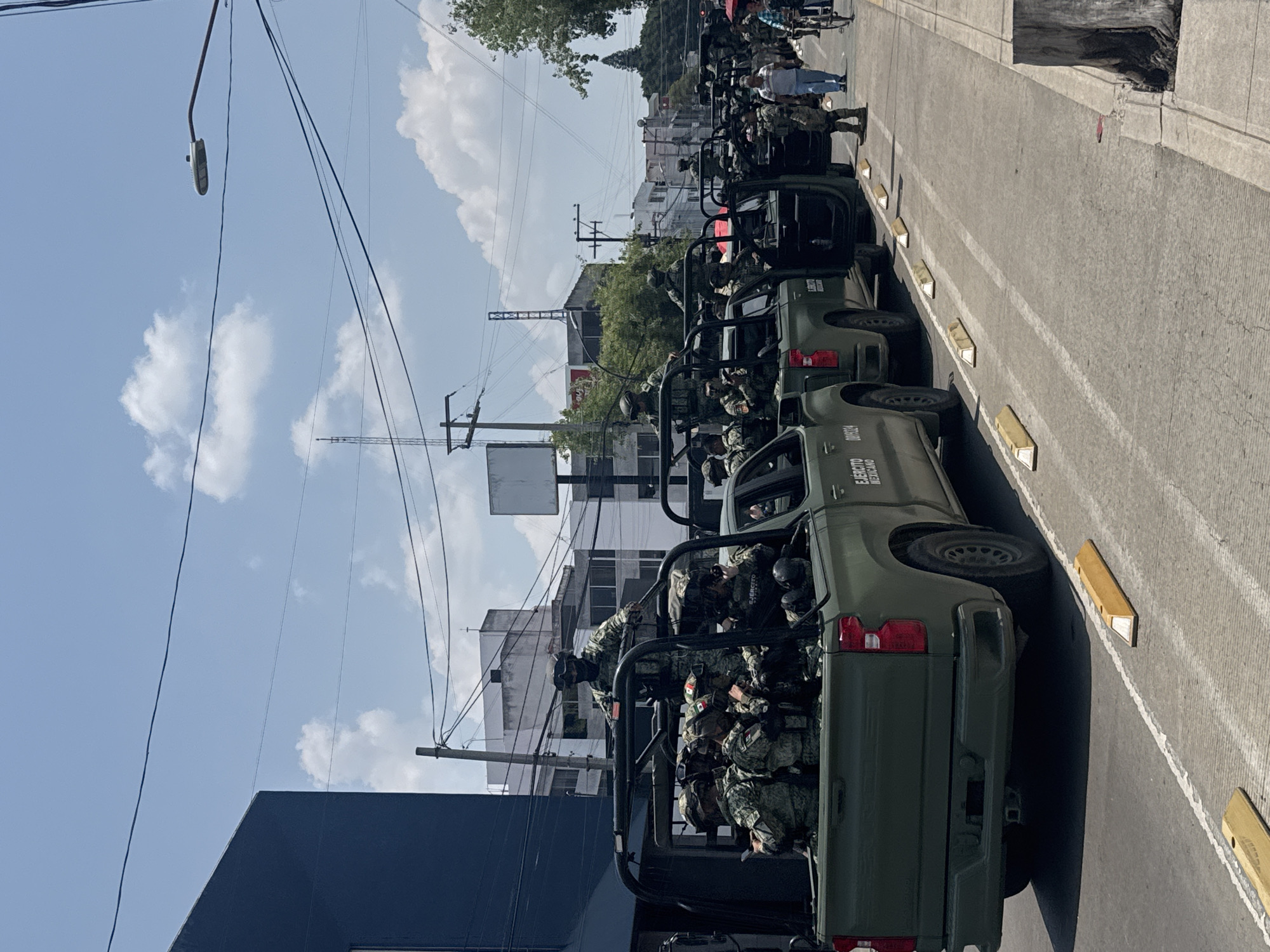 The image shows a street in Puebla, Mexico lined with olive green military pickup trucks. Soldiers in camouflage uniforms and helmets are riding in the beds of the trucks. The vehicles are parked close together along the right side of the road. The road surface has white lane markings and yellow curb markers. Buildings and utility poles are visible in the background. The sky is blue with scattered clouds. Pedestrians are visible further down the street.