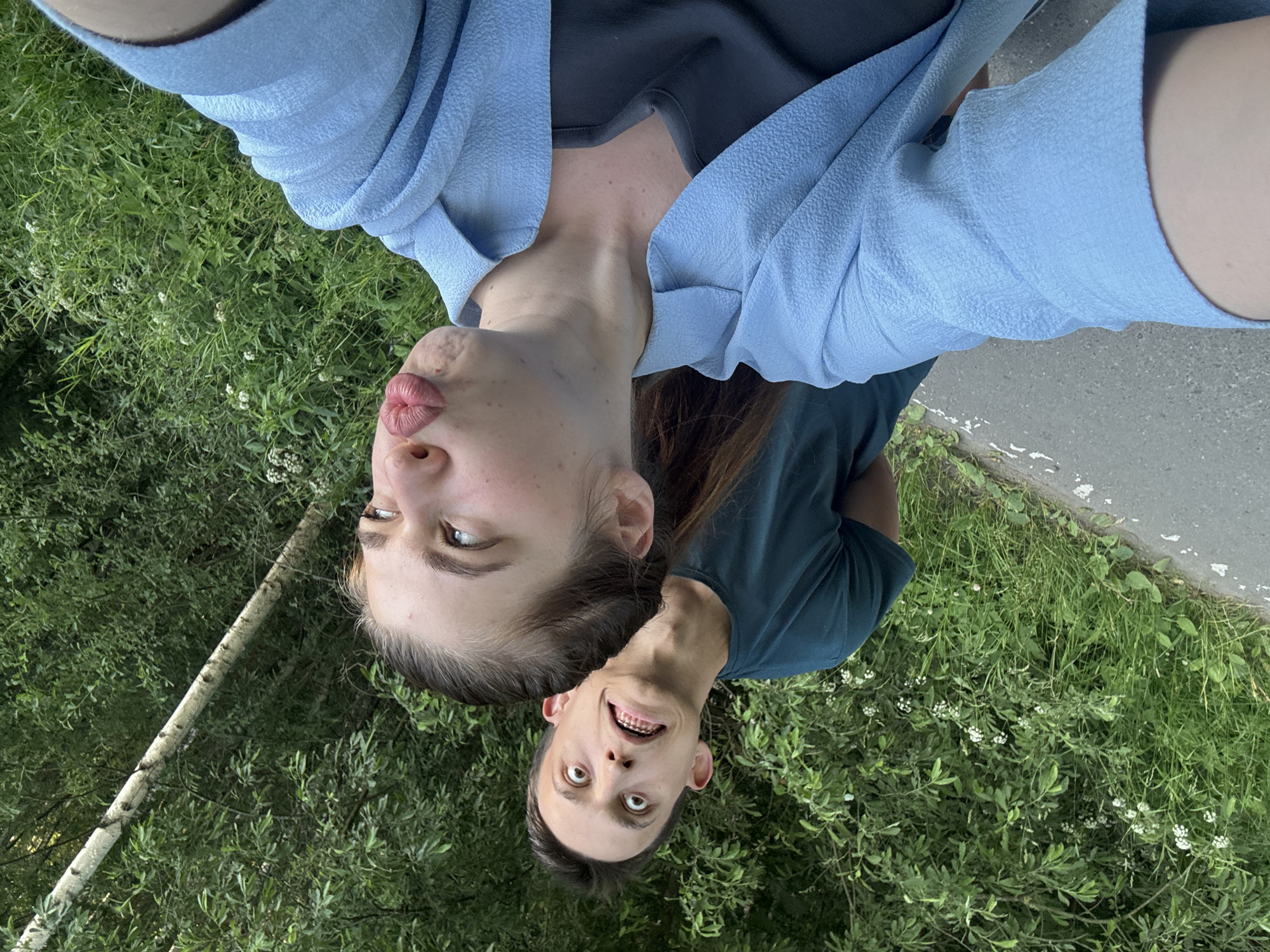 The media file is a slightly blurry, rotating, selfie-style image of two people outdoors, in what appears to be a park or natural area. The foreground shows the faces of a young woman and a young man, both looking directly at the camera. The woman, on the left, is making a kissing face, with her lips pursed and her eyes looking slightly to the side. She is wearing a light blue, textured blouse over a dark grey top. The man, on the right, has a wide-eyed, surprised expression and is grinning, showing his teeth, some of which appear to have braces. He is wearing a dark teal t-shirt.
Behind the subjects, lush green foliage and some white wildflowers are visible, suggesting a warm, possibly spring or summer day. To the right of the man, a grey paved surface, possibly a path or road, is visible. A slender tree trunk with light-colored bark is also present in the background, to the left of the woman. The lighting suggests it is daytime, likely late afternoon or early evening given the softness of the light. The overall mood of the image appears playful and informal. No text is visible in the image. The location context provided is Шевлягино, Russia.