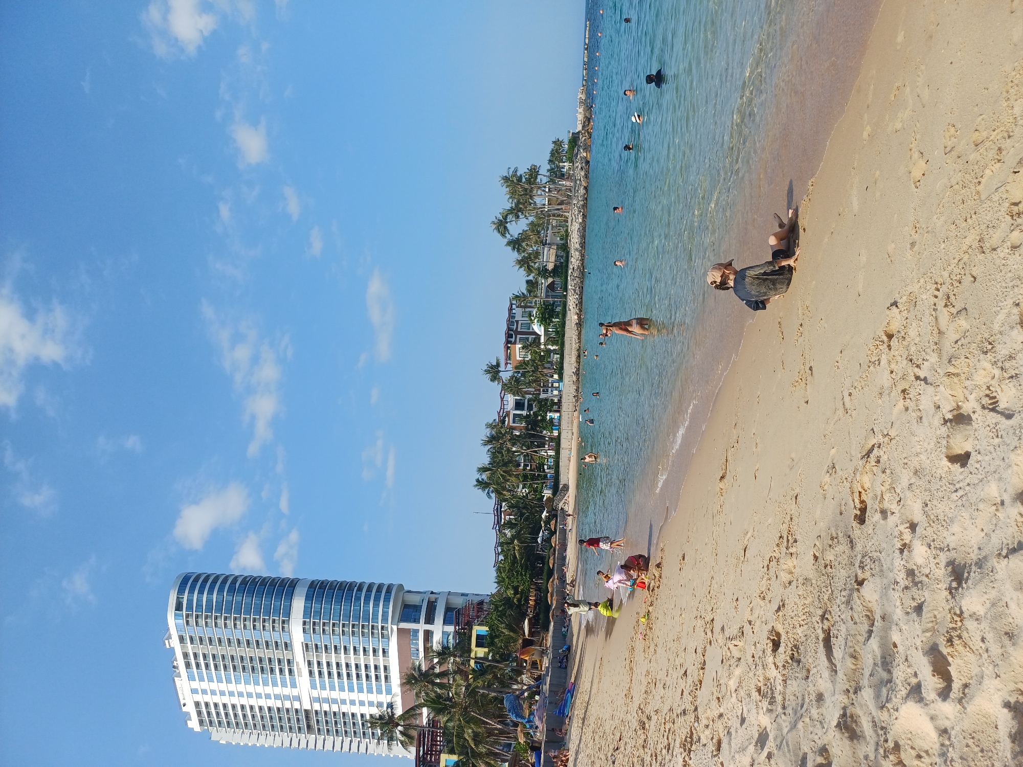 The image depicts a sunny day at a beach in Nha Trang, Vietnam. A tall, modern hotel with a curved facade, likely the InterContinental Nha Trang, dominates the background on the left. Palm trees and other resort buildings line the shore. Numerous people are enjoying the water and sand. In the foreground, a child wearing a cap sits on the wet sand by the edge of the clear blue water. Several other individuals are visible swimming and wading in the sea. The beach is sandy with some footprints and indentations.