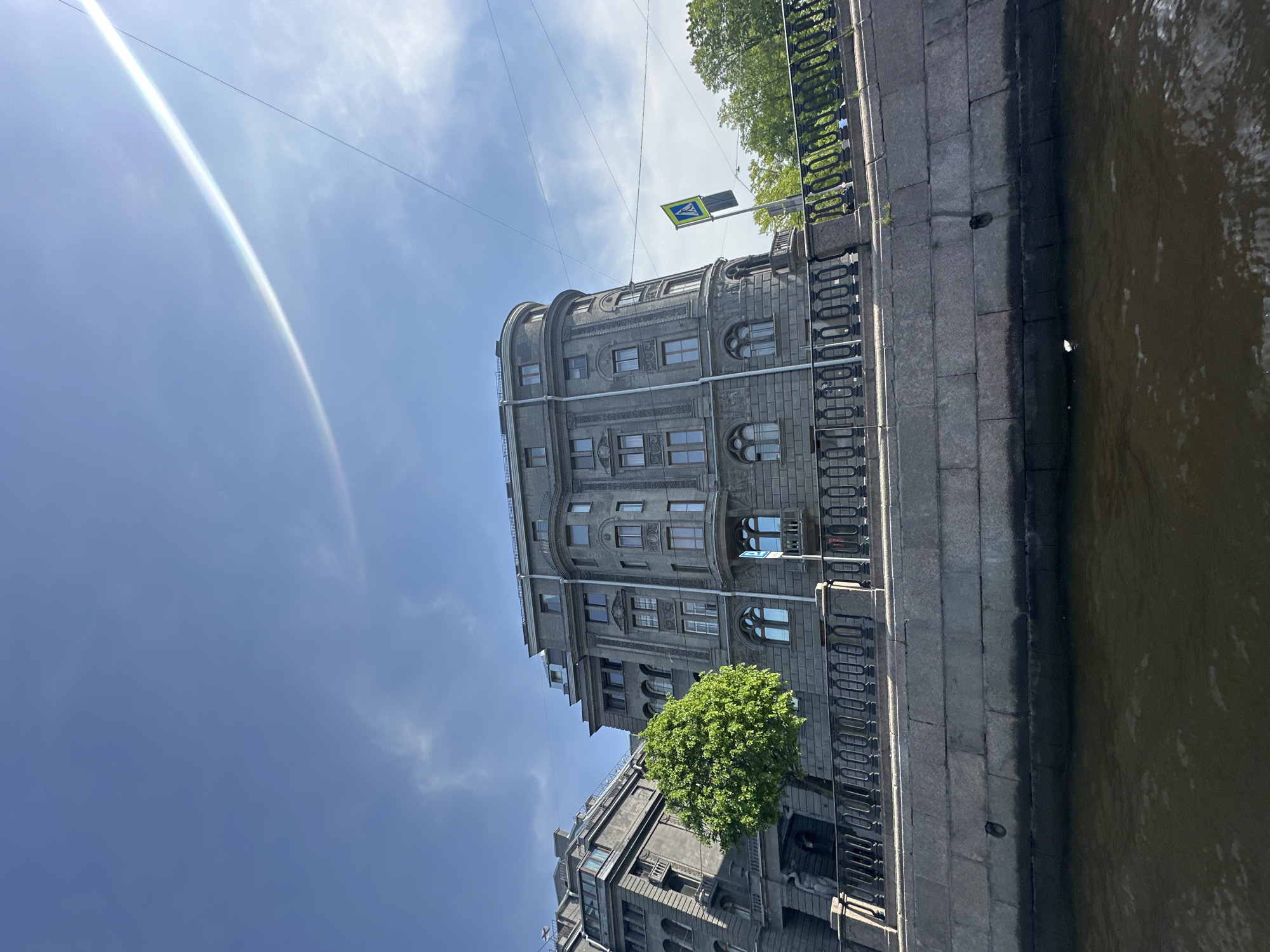 The photograph shows a multi-story building in Saint Petersburg, Russia, situated alongside a canal. The building is constructed of gray stone and features ornate architectural details. The canal is visible to the right of the building, with a stone embankment running along its edge. A pedestrian crossing sign is mounted on a pole in front of the building. The sky is blue with some light clouds.