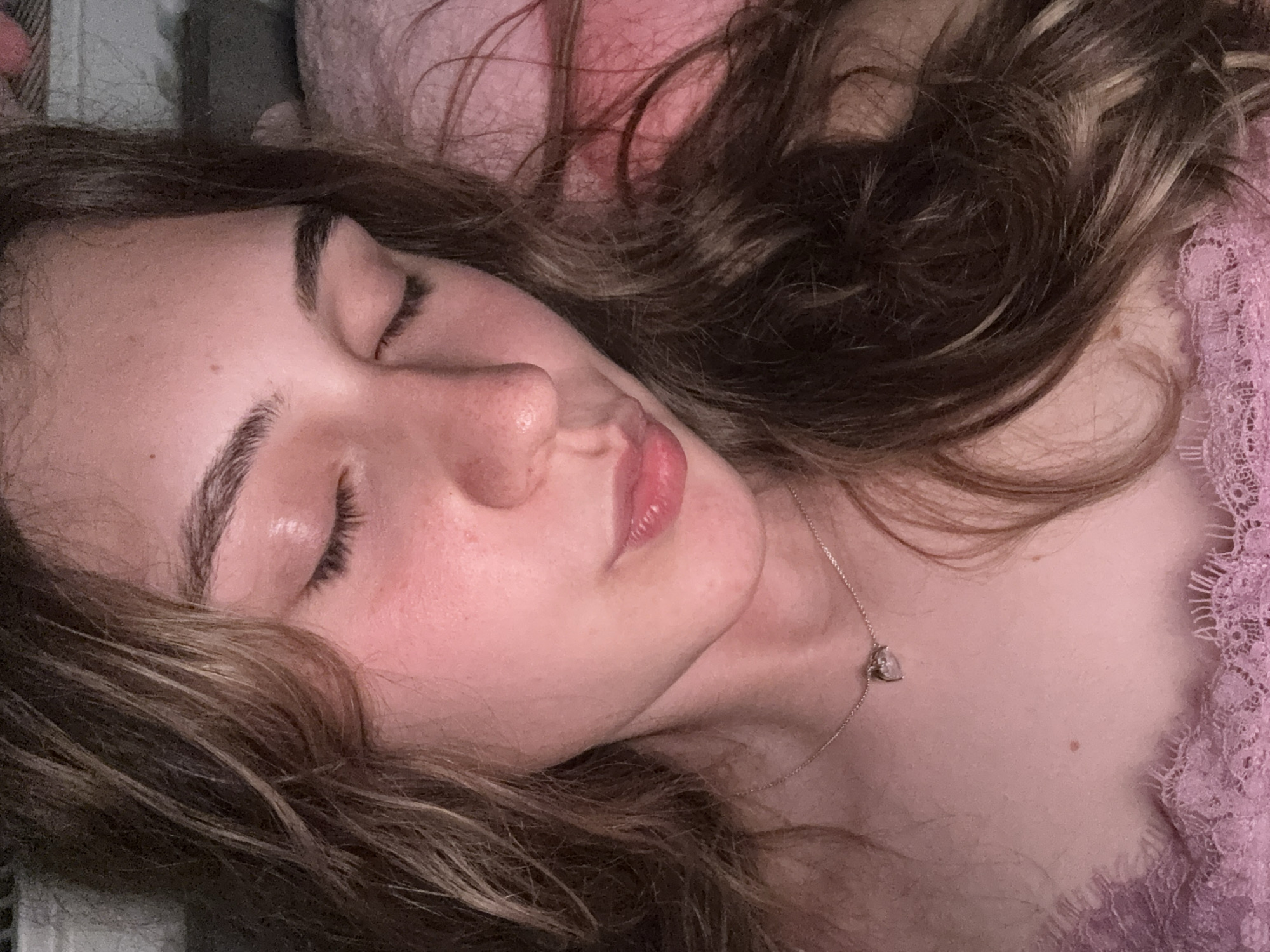 The image is a close-up, indoor, eye-level, personal photograph of a young woman's face from the nose up as she sleeps or rests with her eyes closed. She has brown, wavy hair that frames her face. She is wearing a delicate necklace with a small heart-shaped pendant and a pink lace top. The lighting is soft and warm, casting a pinkish hue across her skin. The background is out of focus but appears to be a soft, pink surface, possibly a pillow or blanket. There are no discernible elements that suggest a specific location in Panagyurishte, Bulgaria.