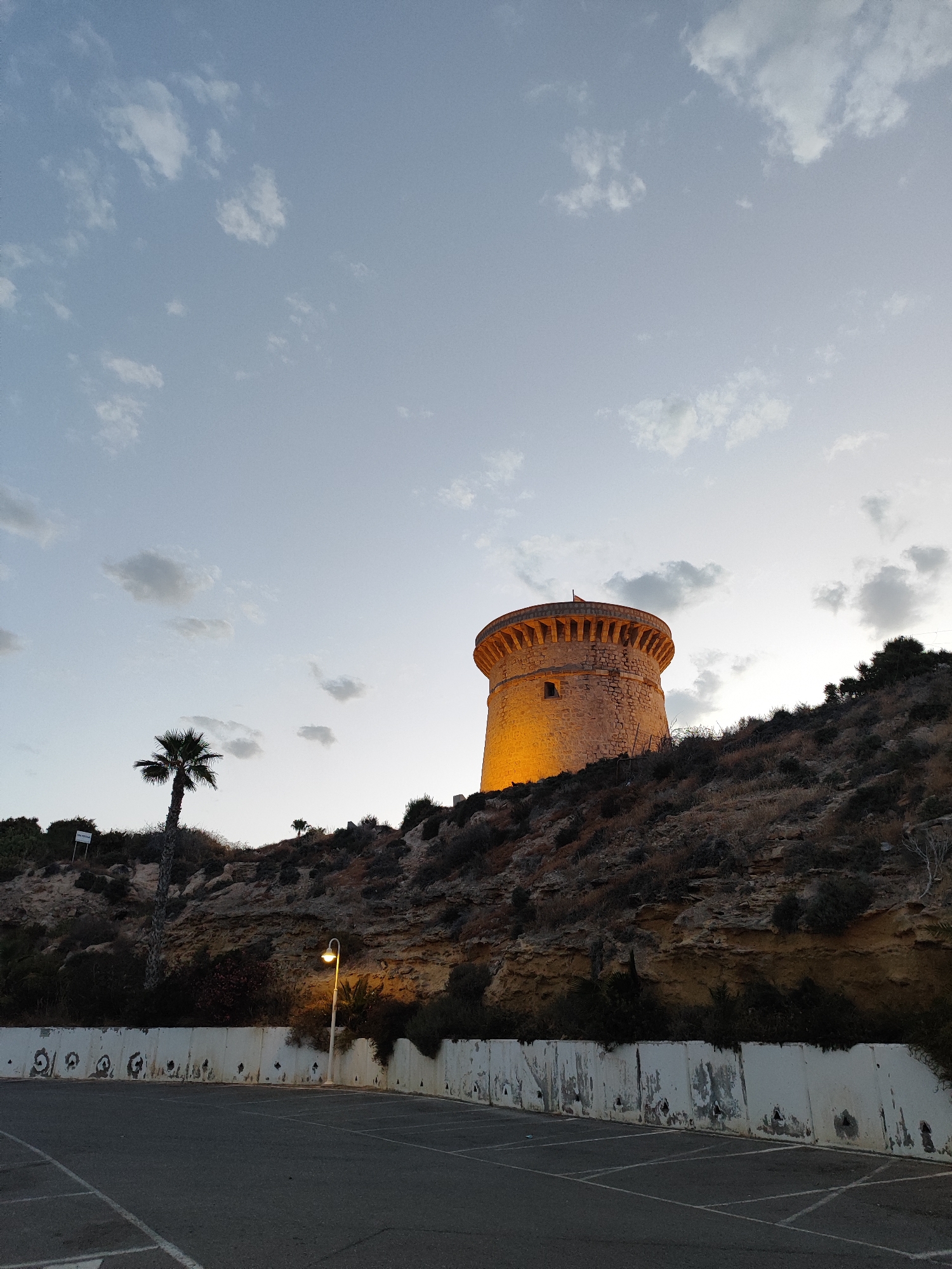 The image depicts the Torre de La Illeta, a historic watchtower, situated in El Campello, Spain. The tower, constructed from stone, stands illuminated by artificial lights, casting a warm glow on its cylindrical form. It is perched on a rocky incline covered with dry brush and sparse vegetation. To the left of the tower, a solitary palm tree stands tall. Below the incline, a concrete wall with weathered markings separates the elevated area from a paved parking lot. The sky above is a pale blue, dotted with scattered clouds, suggesting it is either early morning or late evening. A street lamp with an illuminated bulb is visible near the concrete wall, further enhancing the twilight atmosphere. The scene is devoid of people.