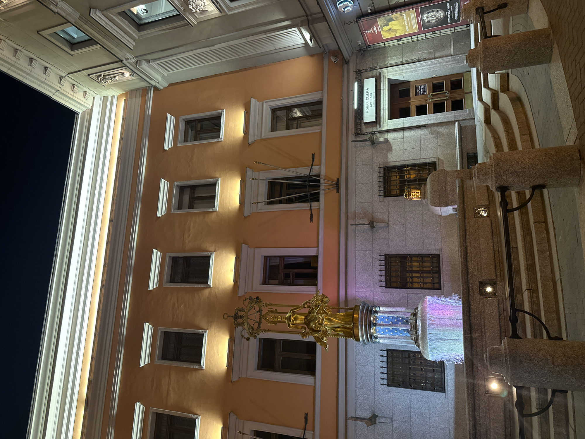 The image captures an exterior view of the "New Stage" Art Cafe at night, located in Moscow, Russia. The facade of the building is illuminated, highlighting its orange walls and architectural details. In the foreground, a golden statue of a woman holding a basket of flowers stands atop a fountain. The fountain features a cylindrical base with a starry pattern and is lit with colorful lights, with water cascading down its sides. Surrounding the fountain are granite steps and railings. To the right of the fountain is the entrance to the "New Stage" Art Cafe, with a sign above the wooden doors. Two posters are displayed on the wall to the right of the entrance. The overall scene is well-lit and showcases the building and its artistic elements in the nighttime urban environment.