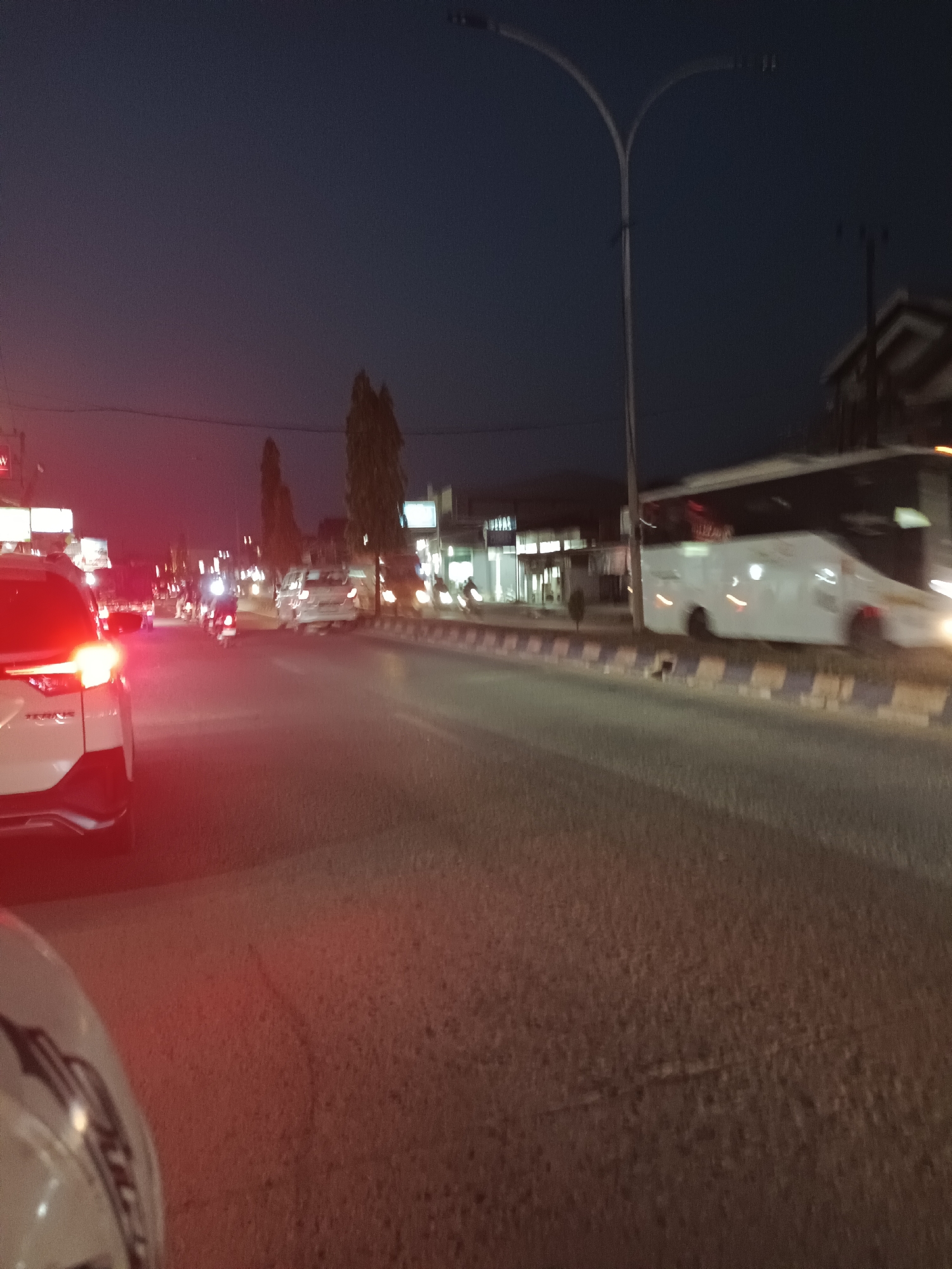 This is a nighttime photograph of a street scene in Teluk Lingga, Indonesia. The image captures a busy road with several vehicles, including cars and motorcycles, moving in the same direction. A large white bus is prominently featured on the right side of the frame, indicating it is either stationary or moving slowly. The street is lined with buildings, some of which are illuminated by lights, suggesting commercial establishments. Trees are visible on the left side of the road, and a street lamp arches over the scene, casting a faint light. The overall atmosphere is that of a typical urban environment at dusk or after dark, with the glow of headlights and streetlights illuminating the asphalt.