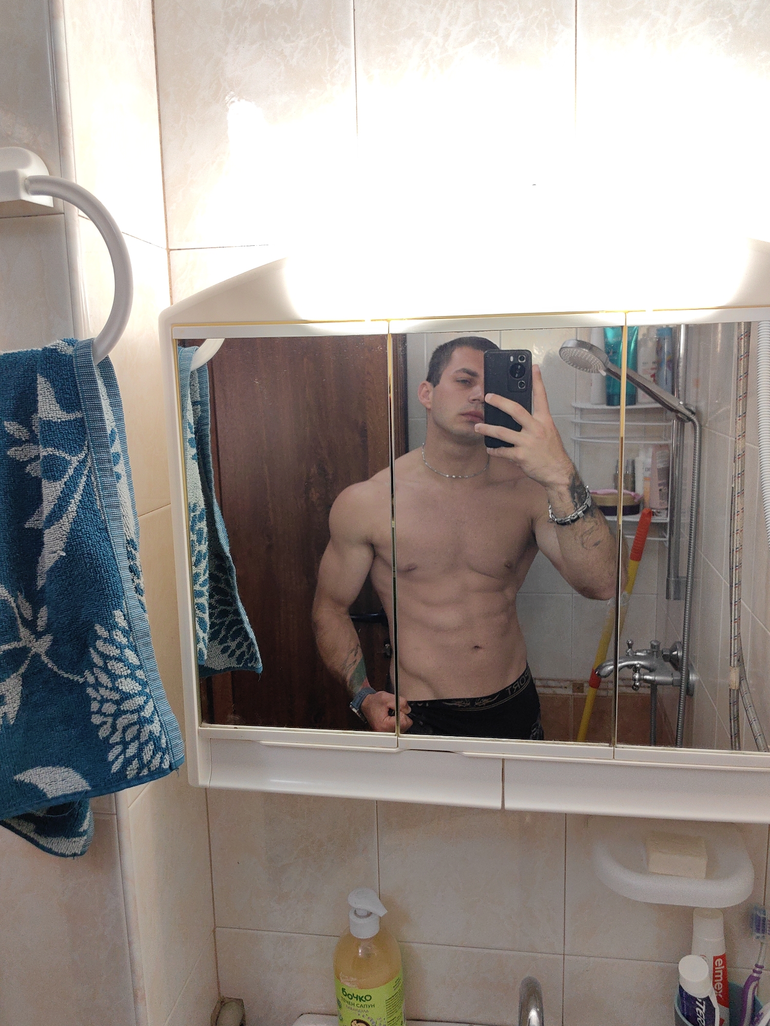 A man is taking a mirror selfie in a bathroom. He is shirtless, wearing black underwear, and has tattoos on his arm. In the reflection, we can see a shower head, various toiletries, and a wooden door. A blue towel with a white floral pattern hangs on a towel ring to the left of the mirror. The bathroom has tiled walls and a tiled floor. The lighting suggests it is daytime. The image is not a clear representation of the city of Varna, Bulgaria, as there are no distinguishable landmarks or context.