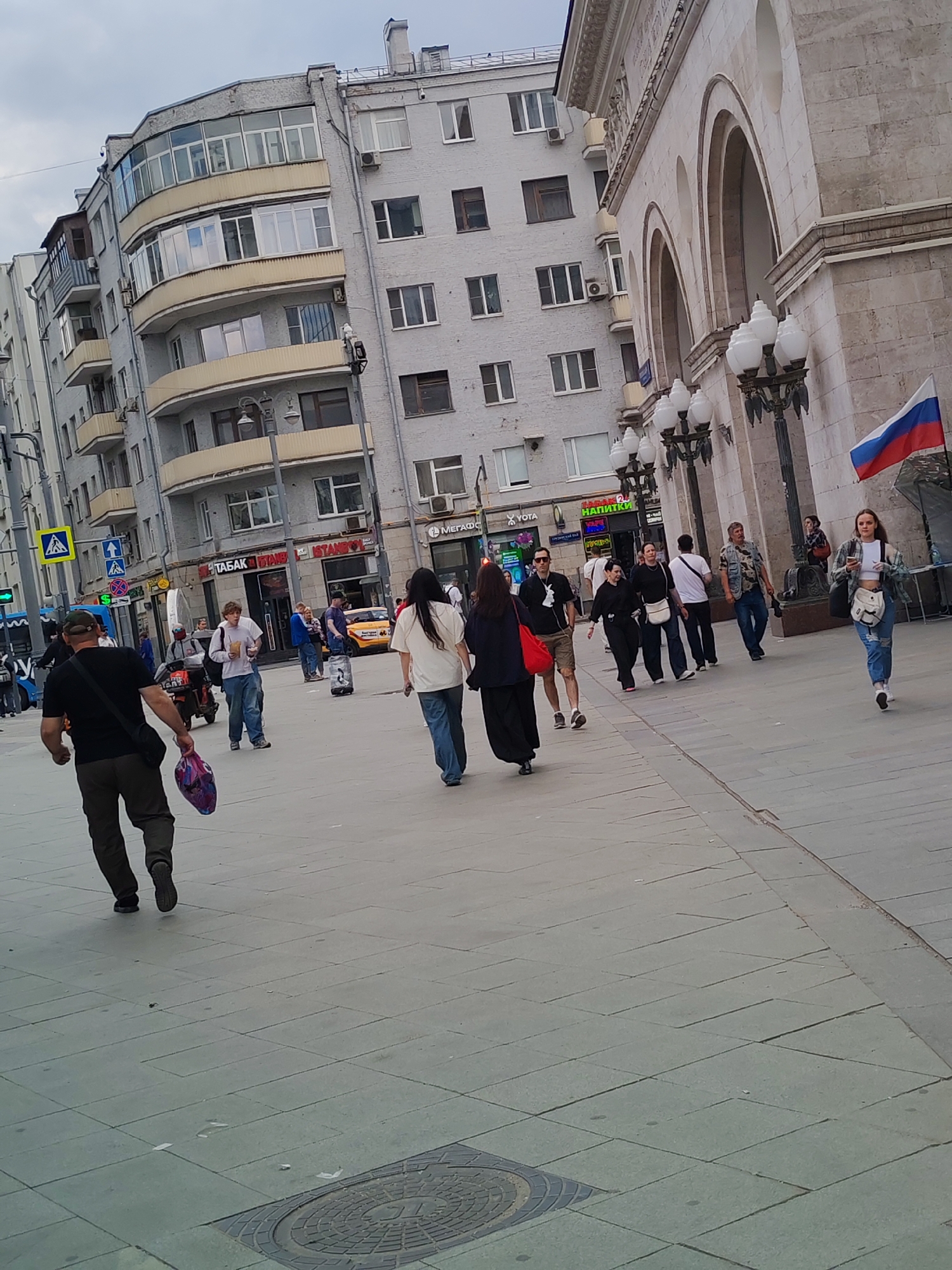 The image depicts a street scene in Moscow, Russia, under an overcast sky. In the foreground, a man wearing a black t-shirt, dark pants, and a cap walks away from the camera, carrying a purple bag. To his left, a group of people walks in the same direction, including a woman in a white top and jeans, and a man in a denim vest and jeans. Further down the street, a young woman in a plaid jacket and ripped jeans walks towards the camera, holding a phone. On the right side of the image, a large, ornate building with arches and columns is visible, featuring a Russian flag waving on a pole. The building appears to be made of light-colored stone. The street is paved with large square tiles, and several lampposts with multiple globes line the sidewalk in front of the building. In the background, multi-story apartment buildings with balconies and windows are seen. Several yellow taxis are parked along the street, and various signs, some in Cyrillic script, are displayed on shopfronts. A traffic light is visible on the left side of the image.