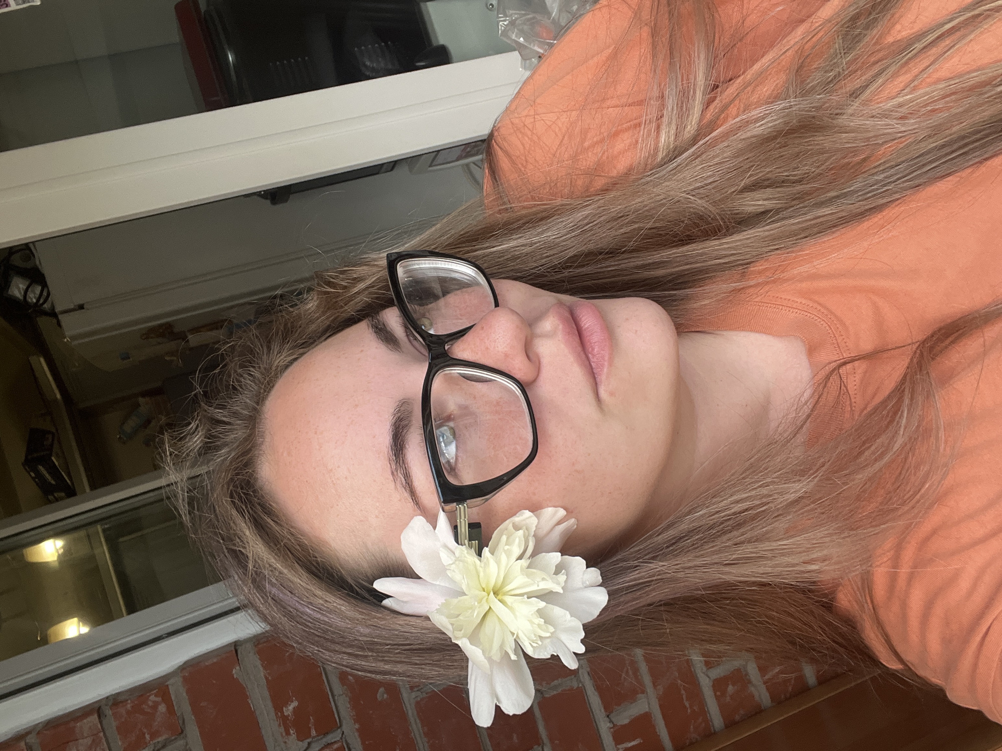 A young woman with long, blonde hair and freckles on her face is positioned in the center of the frame. She is wearing black, cat-eye glasses and an orange t-shirt. A white and yellow flower is clipped to her hair, near her left temple. She is looking off to the right with a neutral expression. Behind her, a red brick wall is visible on the left, and a doorway with white trim is on the right. The background is blurred, but suggests an indoor or semi-outdoor environment with visible reflections in a window. The scene appears to be a casual, close-up portrait. The location is not discernible from the image.
