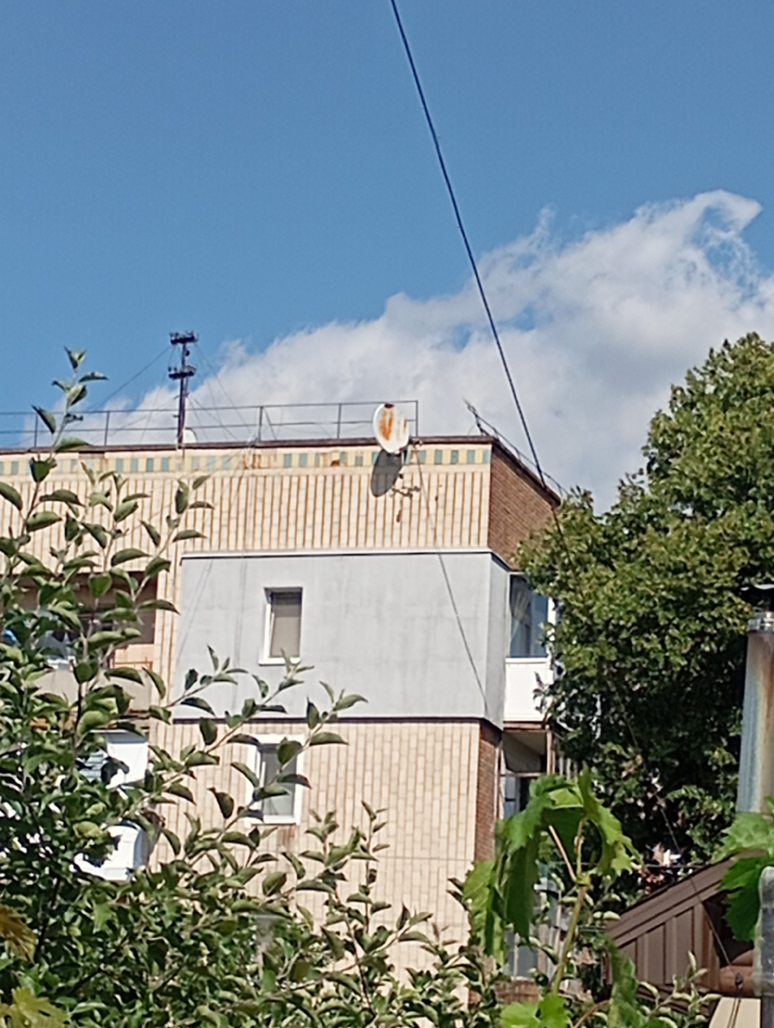 A residential building is seen from below in Sozonivka, Ukraine, on a clear day. The building has a light-colored facade with a beige tiled upper section featuring vertical lines. There are several windows, some of which are dark. Atop the building, a satellite dish with an orange circular marking is visible. A tall, dark pole with horizontal crossbars and supporting wires stands on the rooftop, possibly for antennas. Green foliage from trees and bushes partially obscures the lower portion of the building and extends into the foreground. The sky is blue with scattered white clouds.
