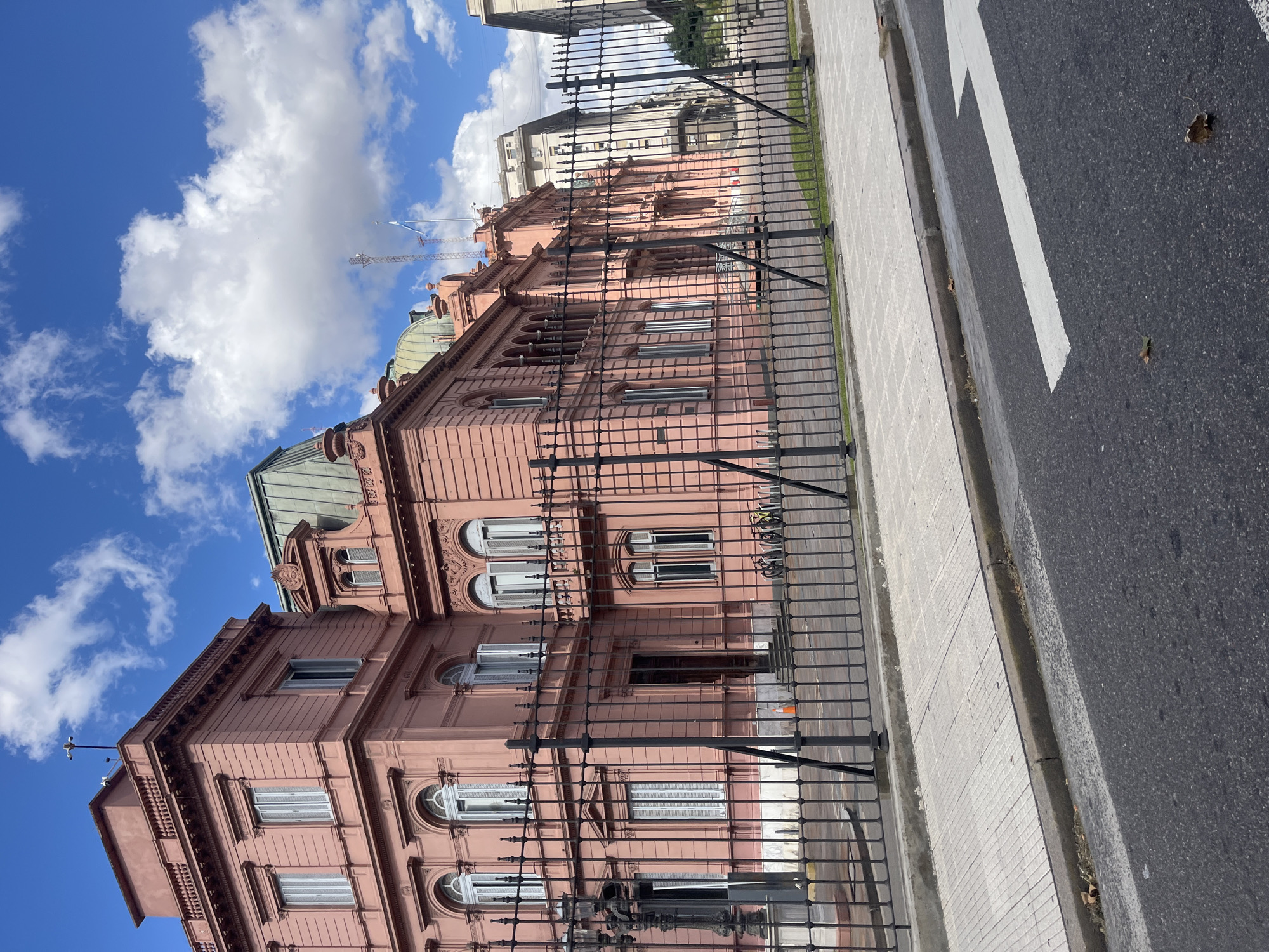 The image shows the Casa Rosada, the presidential palace in Buenos Aires, Argentina. The building is pink with multiple stories and arched windows, and is surrounded by a black wrought iron fence. A sidewalk and street with a white arrow marking are in the foreground. The sky is blue with white clouds. There are other buildings visible in the background. A construction crane is seen in the far background.