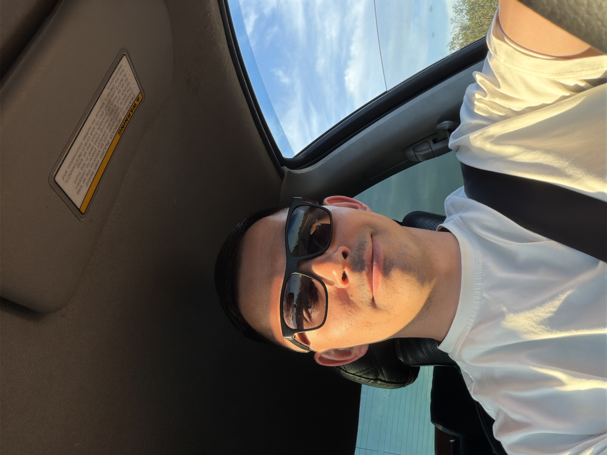 An image taken from a low angle inside a car, likely in Yus'ki, Russia, captures a young man wearing dark sunglasses and a white t-shirt. He appears to be in the driver's seat, looking slightly upwards towards the camera with a neutral to subtly smiling expression. Bright, natural sunlight illuminates the right side of his face and torso, casting soft shadows across his features and clothing, suggesting it is daytime.
The car's interior is visible, including a dark grey headliner, a portion of the sun visor, and a leather car seat headrest. Above, through the car's open sunroof, a clear blue sky with scattered white clouds is visible. A hint of green foliage or trees can be seen in the distance through the car's windows, complementing the sunny weather.
A prominent safety warning label is affixed to the sun visor. The text on the label states: "A WARNING. DEATH OF SERIOUS INJURY can occur. Children 12 and under can be killed by the air bag. The BACK SEAT is the SAFEST place for children. NEVER put a rear facing child seat in the front. Sit as far back as possible from the air bag. ALWAYS use SEAT BELTS and CHILD RESTRAINTS. See the Owner's Manual for further information and explanations."