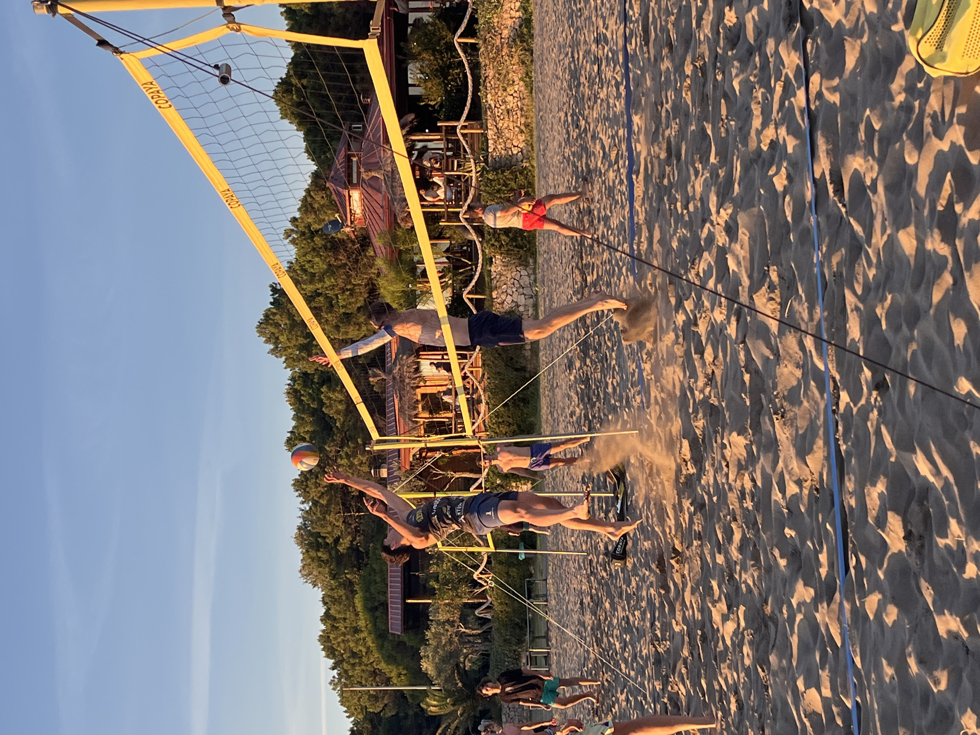 The image depicts a beach volleyball game on a sandy court, possibly located in or near the city of Pistula, Montenegro, as indicated by the context. The setting is outdoors during what appears to be late afternoon or early evening, with the sun casting long shadows across the sand and illuminating the scene with a warm, golden light. A volleyball net, marked with the "COPAYA" logo, stretches across the court.
Two men are actively engaged in a play. One man, wearing a dark tank top and blue shorts, is in mid-air, jumping to hit the volleyball, which is also in the air above the net. The other man, wearing navy blue shorts and no shirt, is also jumping on the opposite side of the net, his arm raised. Other individuals are visible in the background, some watching the game, others appearing to be part of the ongoing match or relaxing nearby.
Behind the volleyball court, there is a restaurant or beach bar structure with a red roof, surrounded by lush green trees. The beach itself is made of fine sand, with some footprints and disturbed sand visible, especially around the players. The sky is clear and blue, with a few faint clouds. The overall atmosphere suggests a casual, recreational sporting event taking place in a coastal environment.