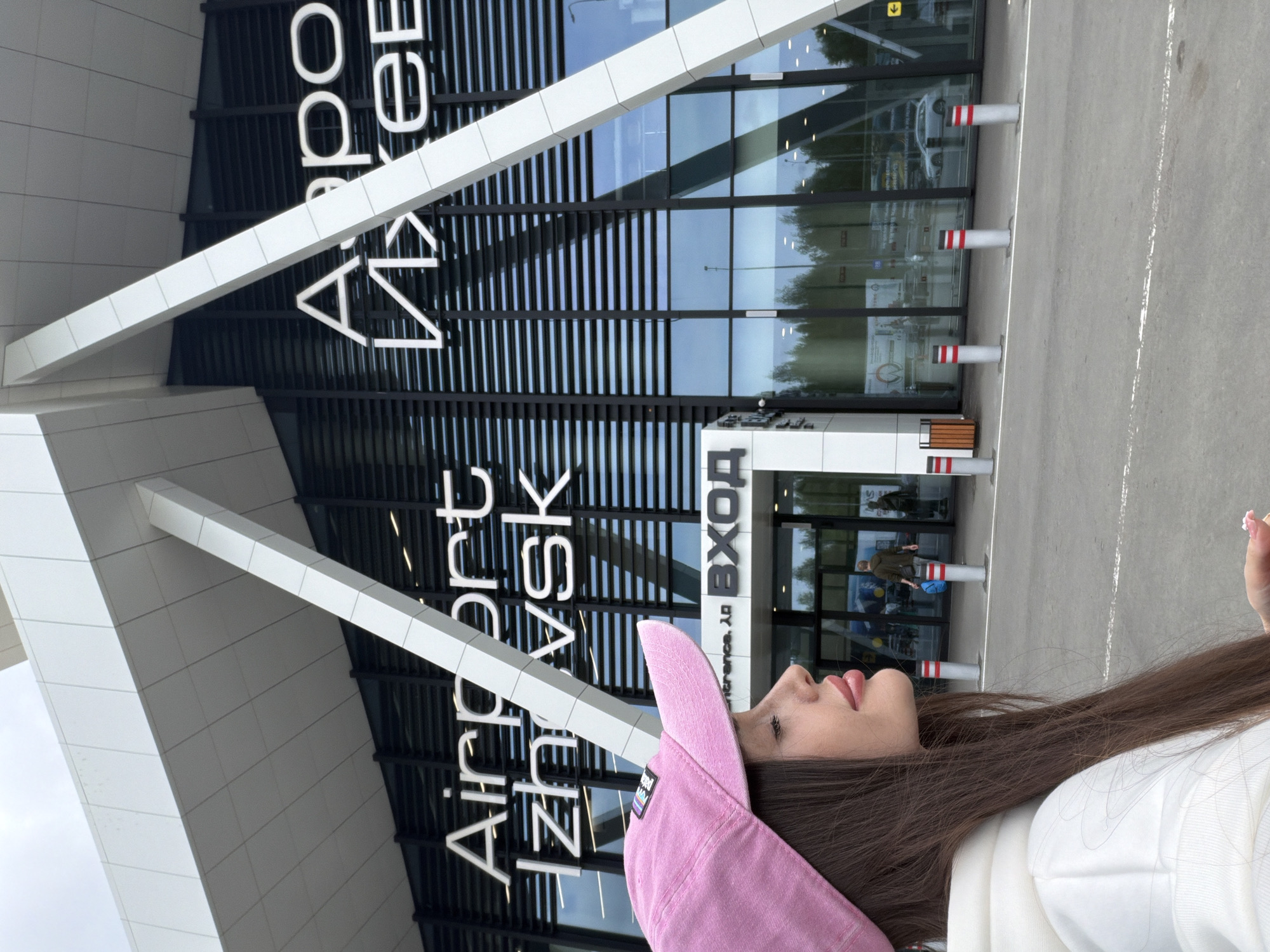 A modern airport terminal building, characterized by large glass facades and white angular architectural elements, stands prominently under an overcast sky. The image is taken from a low angle, showing the building's contemporary design. In the foreground, a young woman is visible in profile, looking towards the right (when oriented correctly). She has long, dark brown hair, wears a pink baseball cap with a small logo, and a white hoodie. Her lips are slightly parted.
The building's exterior features prominent white lettering that spells "Airport Izhevsk". Below "Airport", the word "Izhevsk" is also written in Cyrillic ("Ижевск"). To the right, the Russian word "ВХОД" (Entrance) is visible, with "Entrance. λ口" beneath it. The glass reflects parts of the sky, trees, and what appears to be another person in a brown jacket holding a blue object near the entrance. In front of the terminal, an asphalt ground area with white lines is sectioned off by several red-and-white striped bollards, typical of airport drop-off or short-term parking zones. The scene depicts a moment at Izhevsk Airport, located in Новое Мартьяново, Russia.