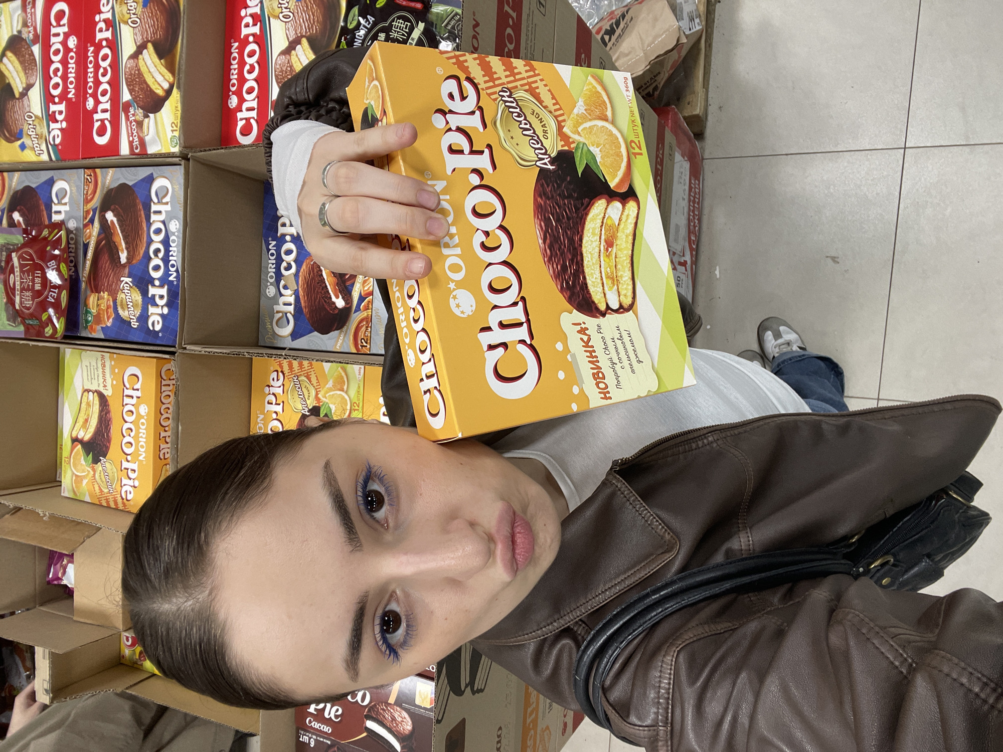 A woman in a brown leather jacket is holding a box of "ORION Choco-Pie" with orange flavor, standing in a store aisle filled with boxes of various Choco-Pie flavors. She is taking a selfie with the product. The packaging is in Russian. The store appears to be in Russia, potentially Moscow, given the presence of Cyrillic text and the widespread availability of Orion Choco-Pie.