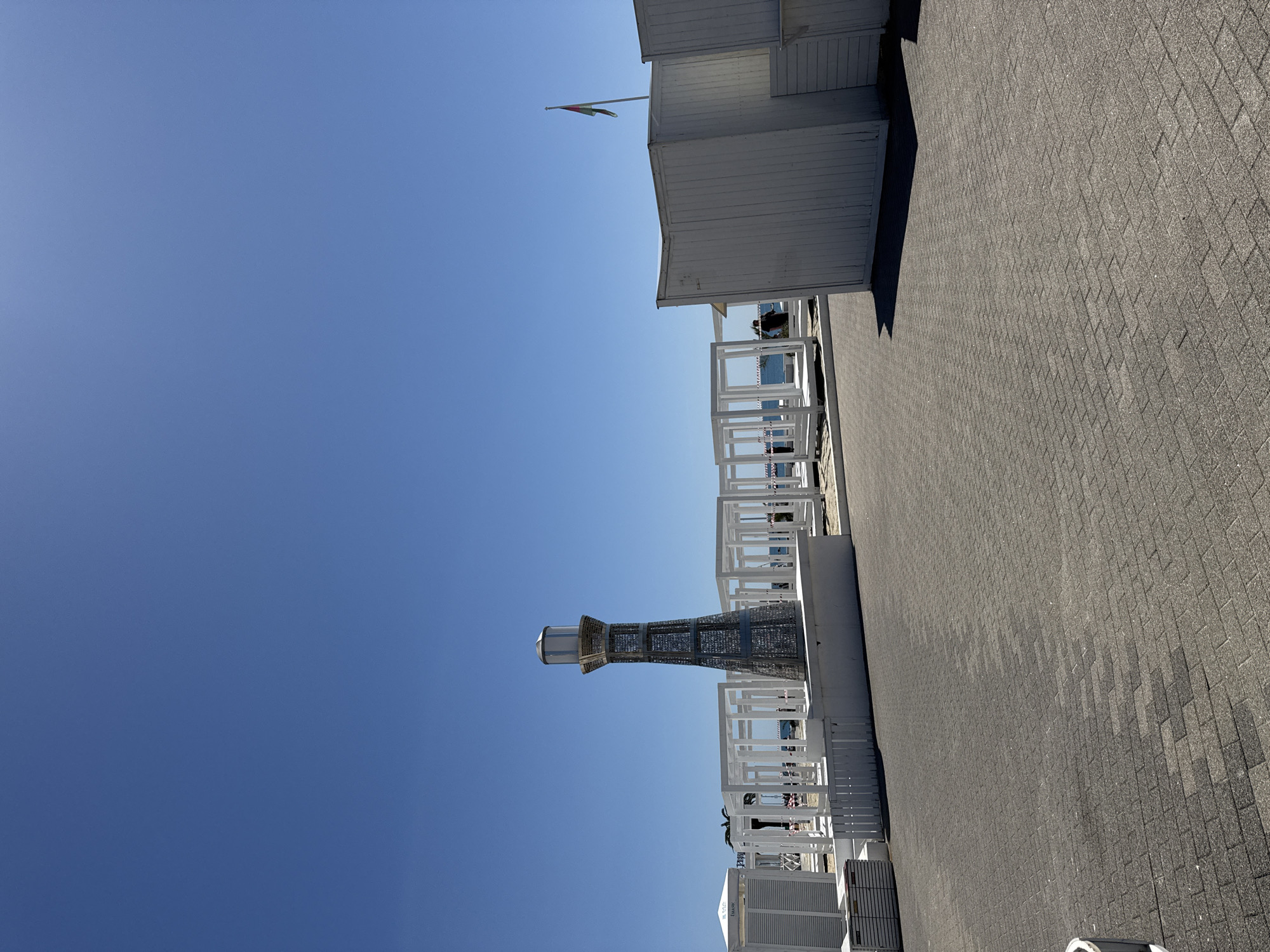 This image captures a bright, sunny outdoor scene, likely a coastal promenade or resort area, with a strong clear blue sky dominating the upper and left portions. The ground on the right and bottom is a greyish-brown paved surface with a distinctive rectangular block pattern.
In the upper right, a simple white building, possibly a booth or small cabin, stands with a flat roof. A flagpole extends from its top, flying a flag with horizontal or vertical stripes of green, white, and red, possibly with an emblem, which could be the flag of Italy. Below this building, and extending into the mid-ground, are several sections of white, slatted railings or balustrades, suggesting an outdoor seating area or terrace. Through these railings, a sliver of distant blue water, likely the sea or ocean, is visible.
A prominent decorative structure resembling a lighthouse stands in the mid-left, constructed from dark, interwoven metal or latticework, topped with a light fixture. A few indistinct figures of people can be seen sparsely placed near the white structures and railings. In the bottom right, white box-like containers or furniture are present, with partially visible text that appears to include "HOTEL" and numbers like "107103". A pink chain or tape is draped over one of the white railings.
The time of day is clearly sunny, with direct sunlight casting harsh shadows, indicating midday or early afternoon. The weather is bright and clear, without any visible clouds. The overall impression is one of leisure and open space by the water.