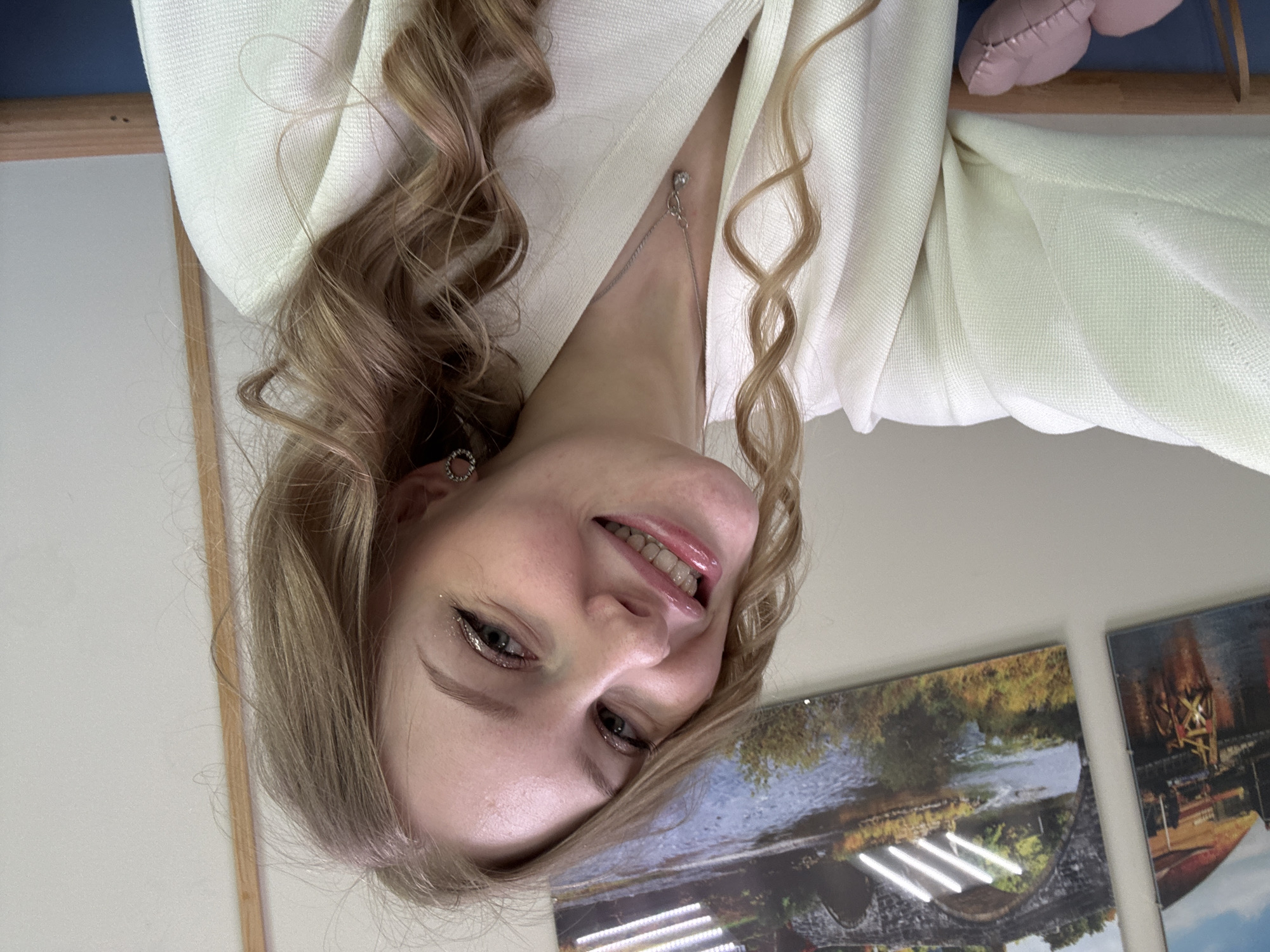 The image is a close-up selfie of a fair-skinned woman with blonde, curly hair, wearing a light-colored jacket or sweater. She is smiling and has light makeup on her eyes and lips. She is wearing earrings and a delicate necklace. Behind her, there are two framed pictures on a wall. One picture shows a river scene, possibly featuring a bridge. The other picture is partially visible and shows what appears to be a building, perhaps a factory. Based on these descriptions, the photo might have been taken in or near the city of Perm, Russia.