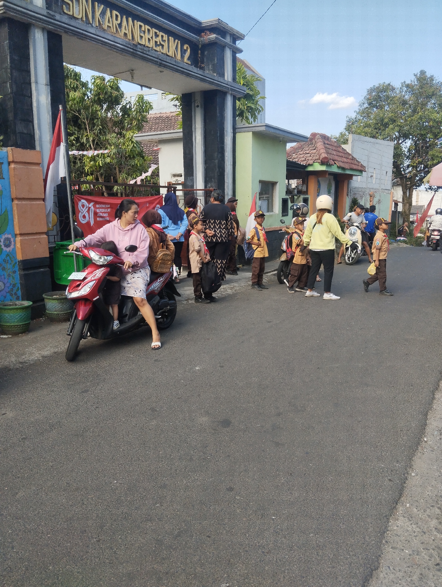 The image depicts the entrance of SDN Karangbesuki 2, a primary school located in Malang, Indonesia. In the foreground, a woman wearing a pink hoodie and camouflage shorts is riding a red scooter with a young child seated in front of her. The scooter has the brand name "Yamaha" visible on its side. To the right of the woman, several children in scout uniforms are standing or walking. One child in the foreground is walking towards the right, carrying a yellow bag. Further back, other adults and children are visible, some on motorcycles, suggesting a busy scene, possibly during school drop-off or pick-up time. A large banner with Indonesian text and the number "81" is displayed prominently near the school entrance. The school gate is decorated with the Indonesian flag and white banners. The environment appears to be a street in front of the school, with trees and buildings visible in the background. The sky is clear and blue with a few clouds.