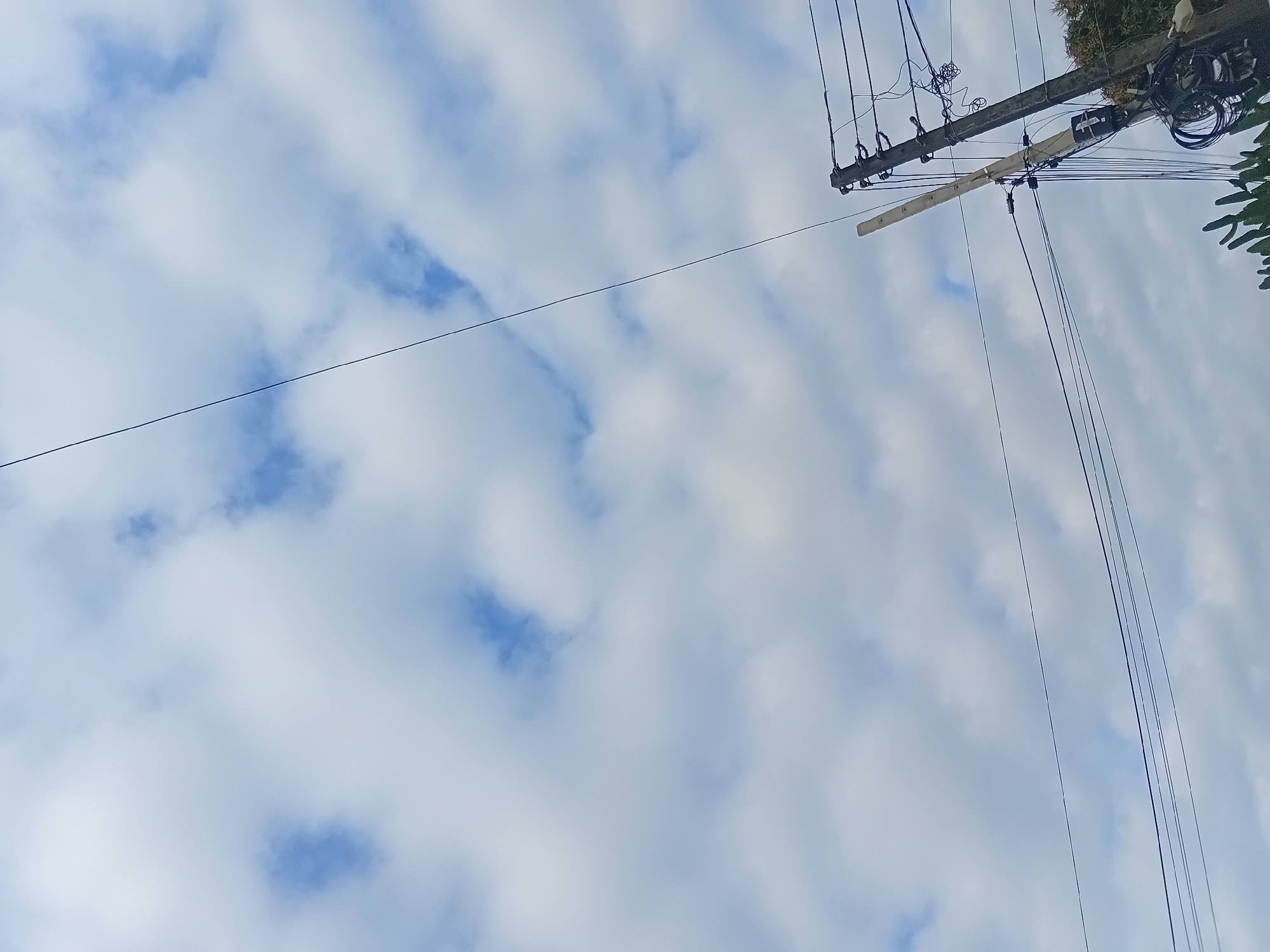 The image is an outdoor, low-angle shot of a cloudy sky with electrical poles and wires in the foreground. The sky is filled with fluffy white clouds and patches of blue. A single dark wire stretches diagonally across the upper half of the frame. On the right side of the image, a dark gray electrical pole stands tall, adorned with numerous wires and various electrical components. Another, lighter-colored pole, possibly an antenna or a communication device, is attached to the main pole. Some greenery, likely from trees or plants, is visible at the bottom edge of the frame. The overall scene suggests an urban or semi-urban environment with overhead electrical infrastructure.