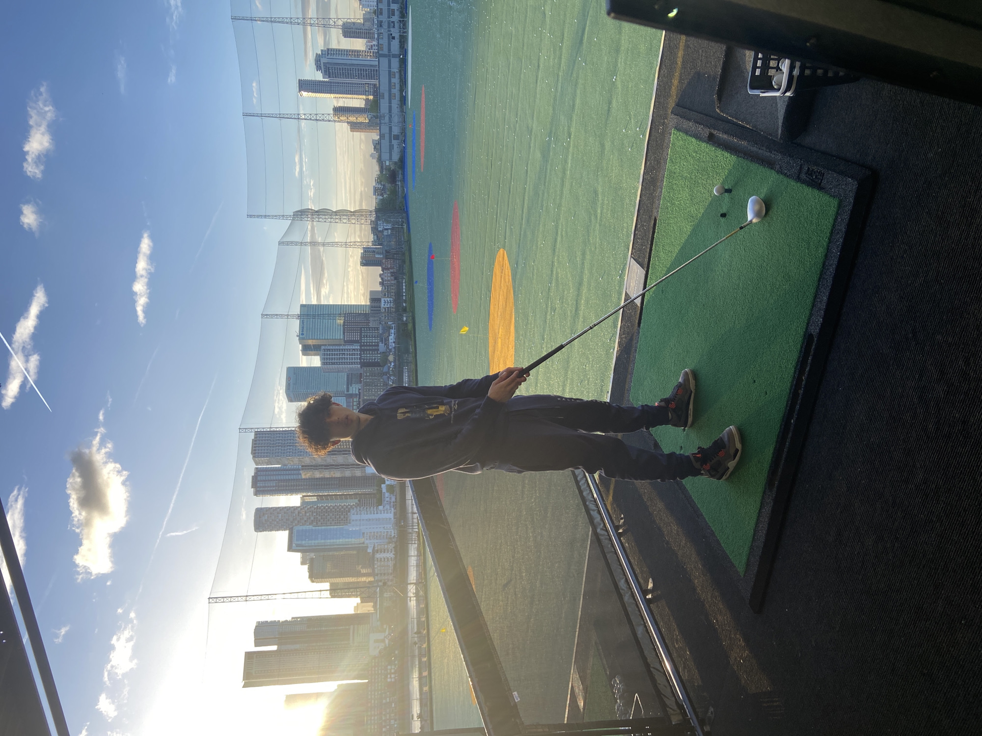 The image depicts a young man at an outdoor golf driving range, situated on an elevated platform overlooking a city skyline. He is positioned on a green artificial turf mat, holding a golf club, with a golf ball ready on a tee beside him. A white driver headcover and another golf ball are also on the mat, and a golf ball dispenser is visible to the right. The man wears a dark hoodie with "RALPH LAUREN" visible on the front, dark sweatpants, and athletic shoes.
The scene captures the man preparing to take a swing, focused on the ball. The driving range extends into a large green area with colorful target markers (red, blue, yellow) and is enclosed by a tall netting. Beyond the netting, a dense array of modern skyscrapers and high-rise buildings forms the urban background, characteristic of London.
The time of day appears to be late afternoon or early evening, indicated by the warm, golden light casting long shadows and creating a bright glow on the horizon and building surfaces. The sky is clear blue with sparse white clouds and a visible airplane contrail. The overall impression is an active leisure setting within a vibrant urban environment.