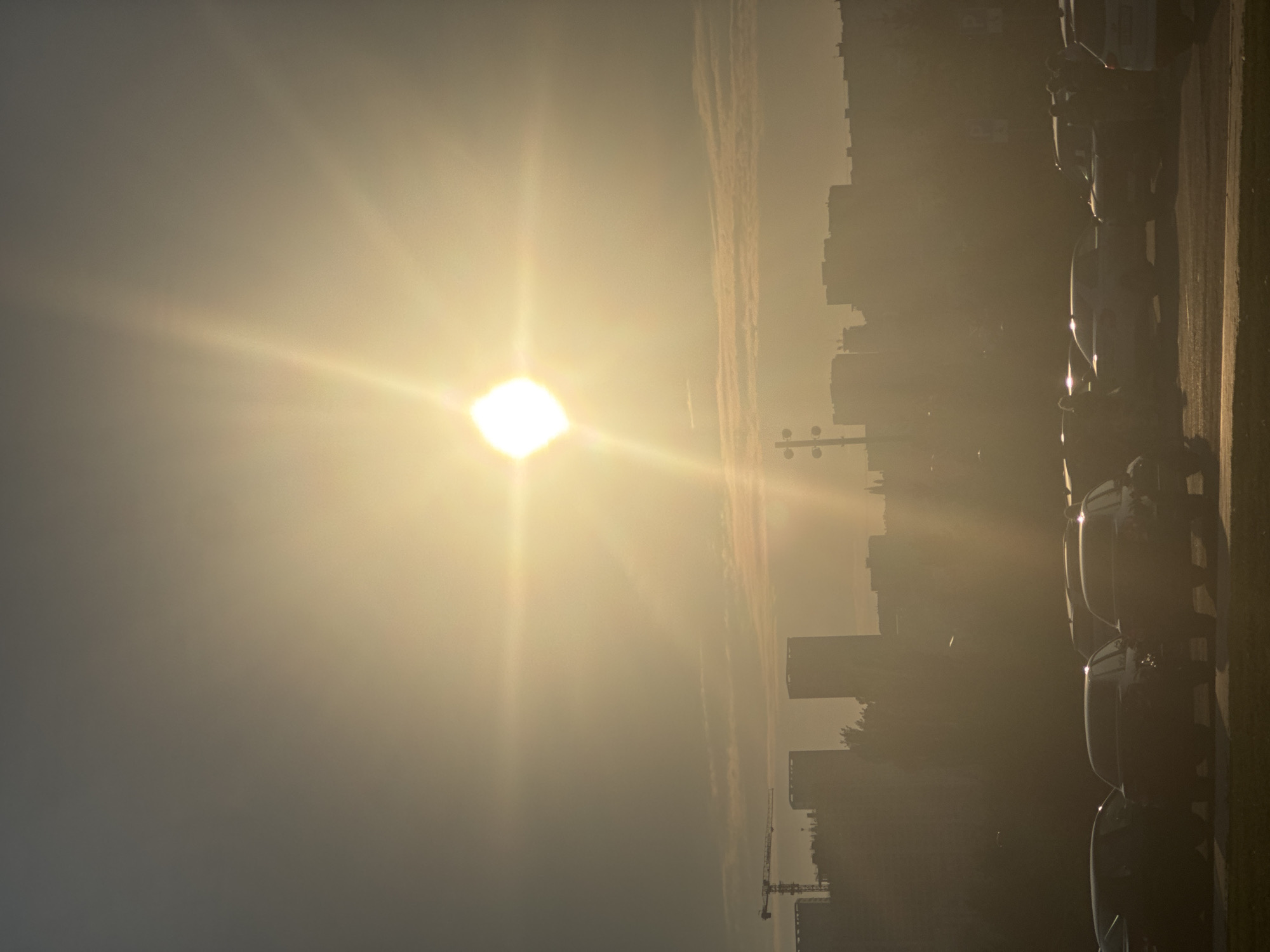The image shows a hazy, sun-drenched cityscape, likely Ufa, Russia, during what appears to be sunset or sunrise. The sun, a bright, glowing orb, dominates the upper half of the frame, casting a strong lens flare and radiating bright beams across the sky. Below the sun, a silhouetted skyline of buildings and construction cranes rises against the luminous haze. A row of parked cars, mostly dark-colored sedans and SUVs, occupies the foreground, their forms indistinct due to the low light and atmospheric conditions. The overall impression is one of a warm, diffused light engulfing an urban environment.