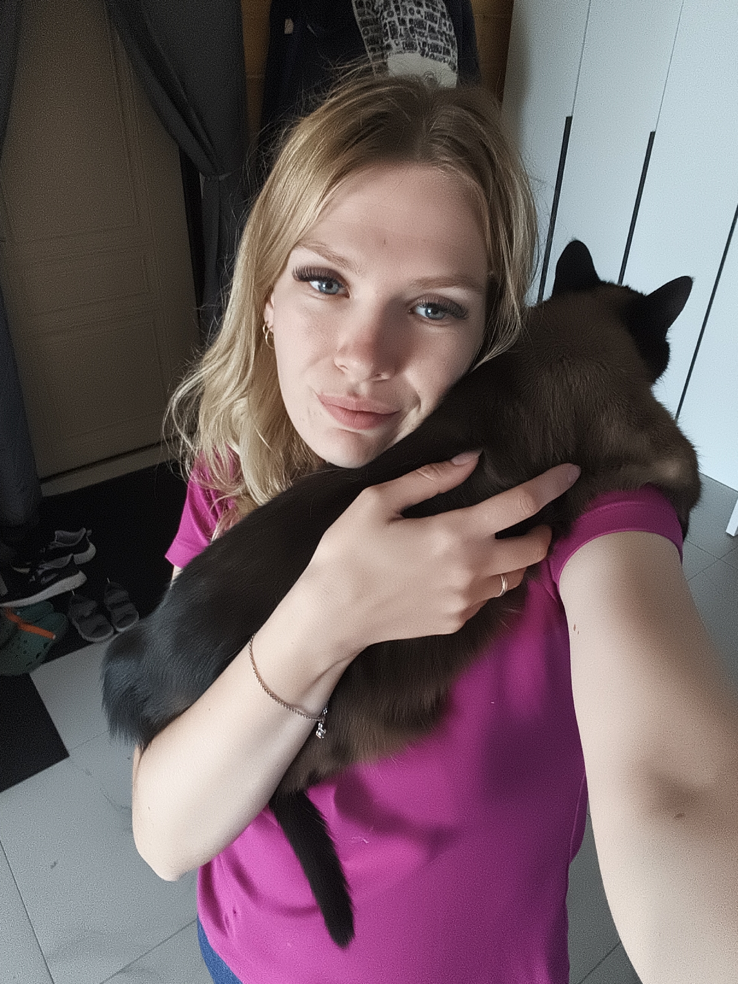 A young woman with blonde hair and blue eyes is holding a dark brown cat in her arms. She is wearing a pink t-shirt and a silver bracelet. The cat is nestled against her shoulder, with its tail hanging down. The background appears to be an indoor setting, with a door and a closet visible. There are also a few pairs of shoes on the floor in the background. The overall impression is one of a casual, domestic scene.