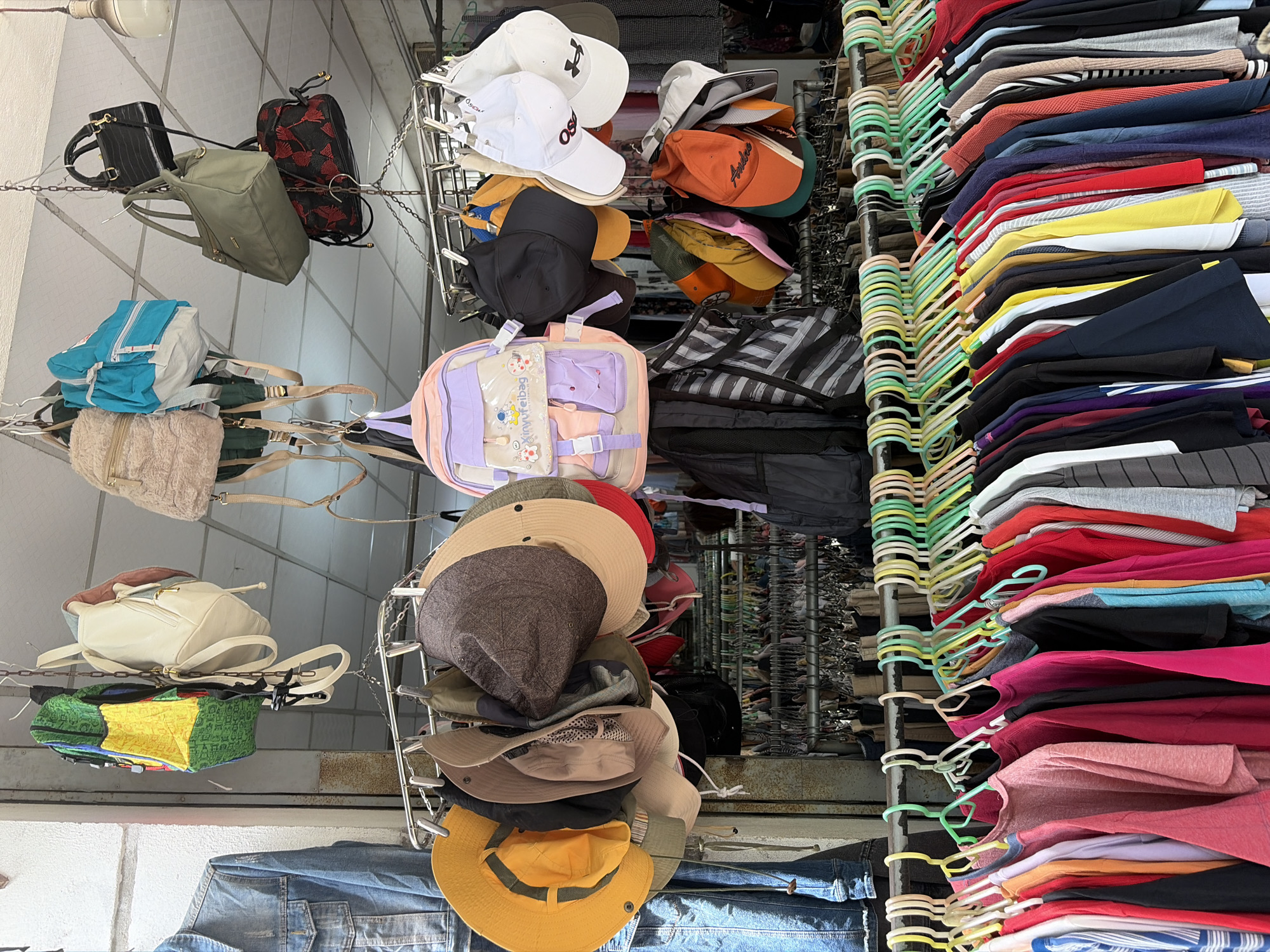 This image depicts a street-side stall in Đà Nẵng, Vietnam, selling a variety of clothing and accessories. The stall features a rack of colorful t-shirts and other garments hanging on hangers, with a selection of hats and bags displayed above. The items appear to be for sale, suggesting a retail environment. The backdrop shows a building facade and overhead structures, typical of an urban market setting. No people are visible in the frame.