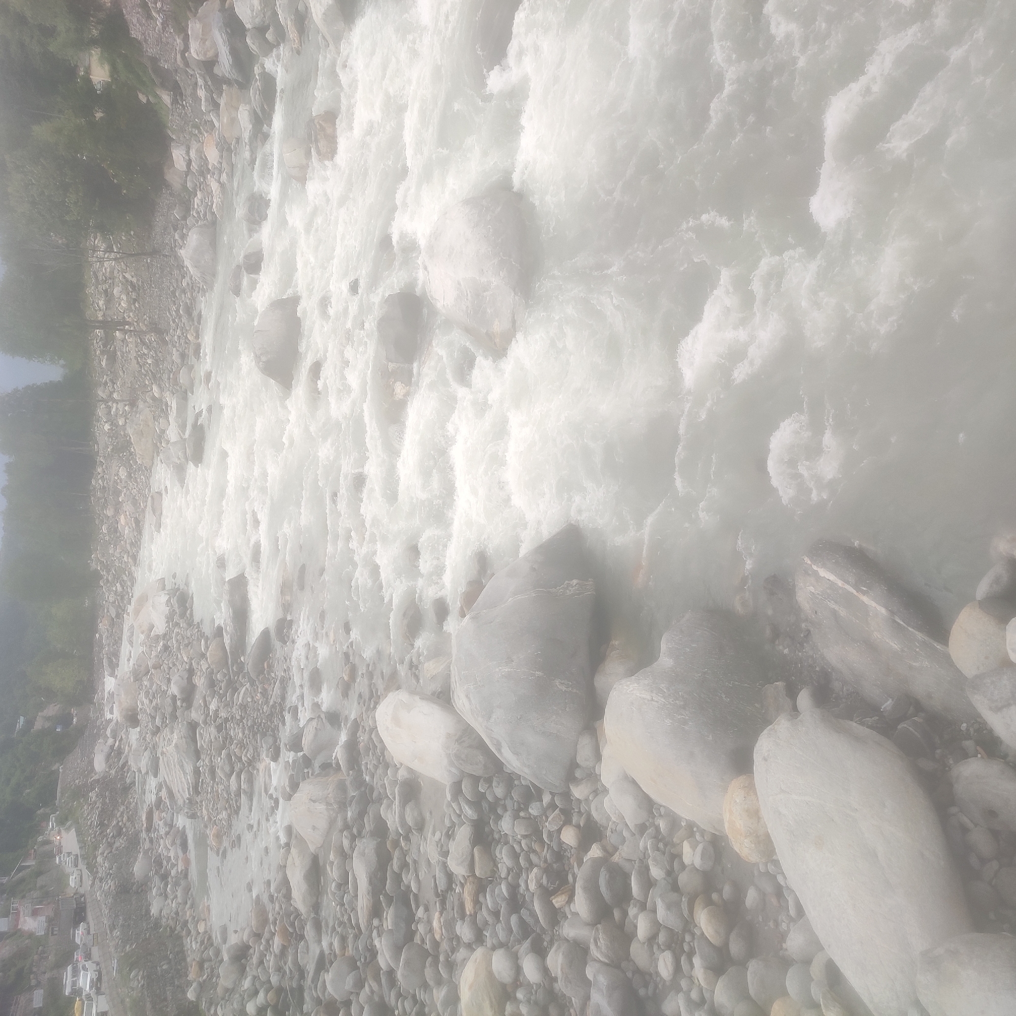 The image depicts a wide, fast-flowing river in Kalath, India, characterized by its turbulent, whitewater surface and numerous large, smooth rocks on its banks and within the riverbed. The river's water appears milky or sediment-laden, suggesting it may be a glacial river or carrying a significant amount of silt. The surrounding environment is mountainous, with steep, rocky slopes visible in the background, partially obscured by mist or fog. Trees and vegetation can be seen on the upper slopes, and a road with several vehicles, including cars and a truck, runs along the edge of the riverbank in the upper left portion of the image. The overall impression is of a natural, wild landscape with human infrastructure present along its edge.