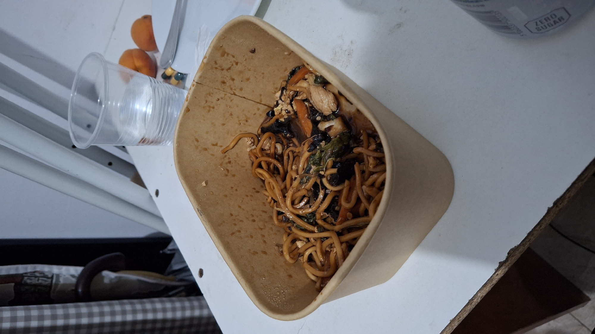 A carton of noodles with chicken and vegetables sits on a white table. Next to the carton is a plastic cup filled with water, two apricots, and a fork on a plate. In the background, there is a patterned curtain and some white supports. The scene appears to be set indoors. The location is Nesebar, Bulgaria.