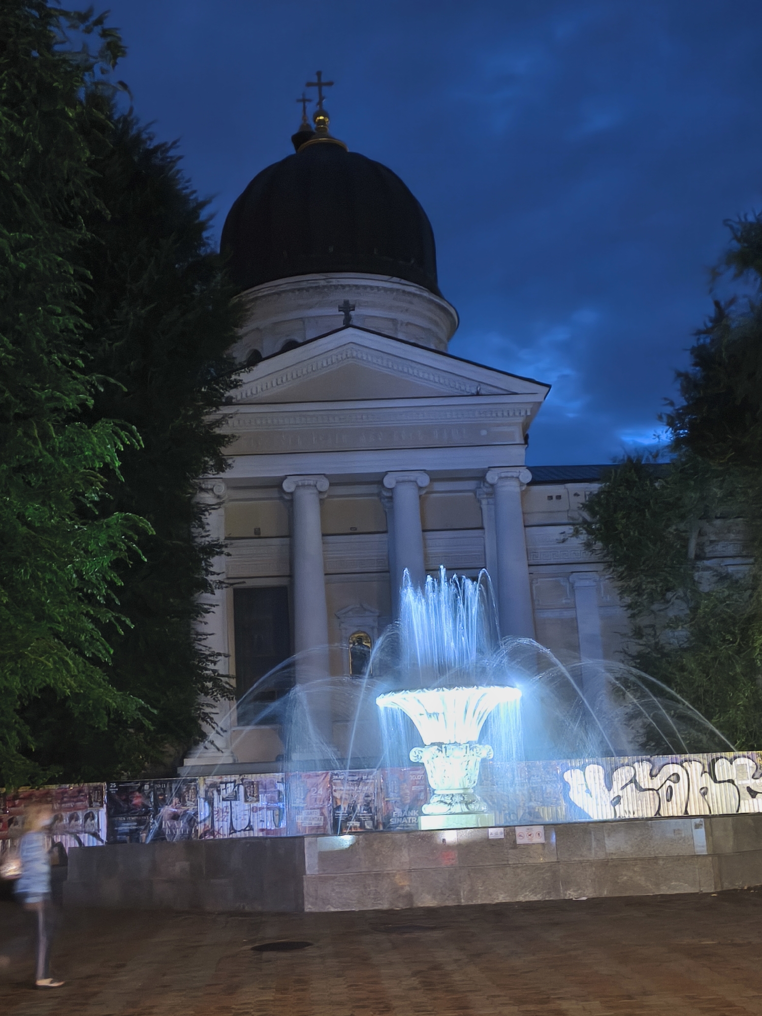 The image captures a night scene in Odesa, Ukraine, featuring a prominent fountain in the foreground illuminated with a bright, bluish-white light. The fountain's water jets spray upwards and outwards, creating a dynamic visual. Behind the fountain, a large building with classical architecture is visible. This building has a dark dome topped with crosses and is supported by several large white columns. The facade of the building is adorned with decorative elements. A low wall surrounds the fountain, and this wall is covered with various posters and graffiti. To the left and right of the central scene, dark silhouettes of trees frame the view. A blurry figure of a person, appearing to be walking, is captured on the left side of the frame, indicating movement and a sense of the urban environment at night. The sky above is a deep blue, suggesting it is dusk or nighttime. The overall atmosphere is one of a public space at night, with the fountain serving as a central illuminated feature.