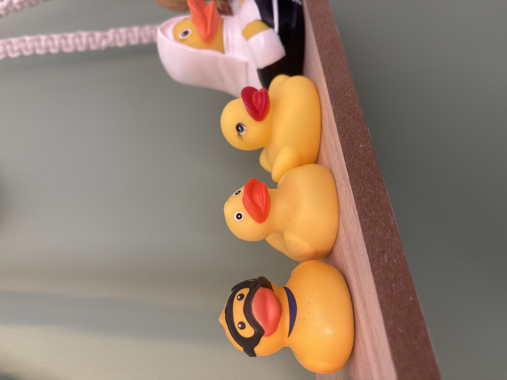 The image displays a close-up, rotated view of five rubber ducks arranged on a light brown wooden shelf or ledge. The background is a plain, soft grey-green wall.
The ducks are lined up vertically in the rotated orientation of the photo. The topmost visible duck, partially obscured, is yellow with an orange beak and appears to be inside a white, open container. Behind it, a yellow rubber duck wears what looks like a white tuxedo or formal uniform with black accents, also sitting within the white container.
Below these, a yellow rubber duck features a red heart-shaped ornament on its head and has painted blue eyes with eyelashes. Next in line is another standard yellow rubber duck with a plain orange beak and simple black dot eyes. The bottommost duck is also yellow but sports a black mask-like design around its eyes and a blue collar, giving it the appearance of a character like a superhero or bandit, and has an orange beak.
To the left of the ducks, a blurred white braided or macrame cord is partially visible against the grey-green background. The ducks are static, displayed as a collection. There is no activity taking place, and no visible text in the image.