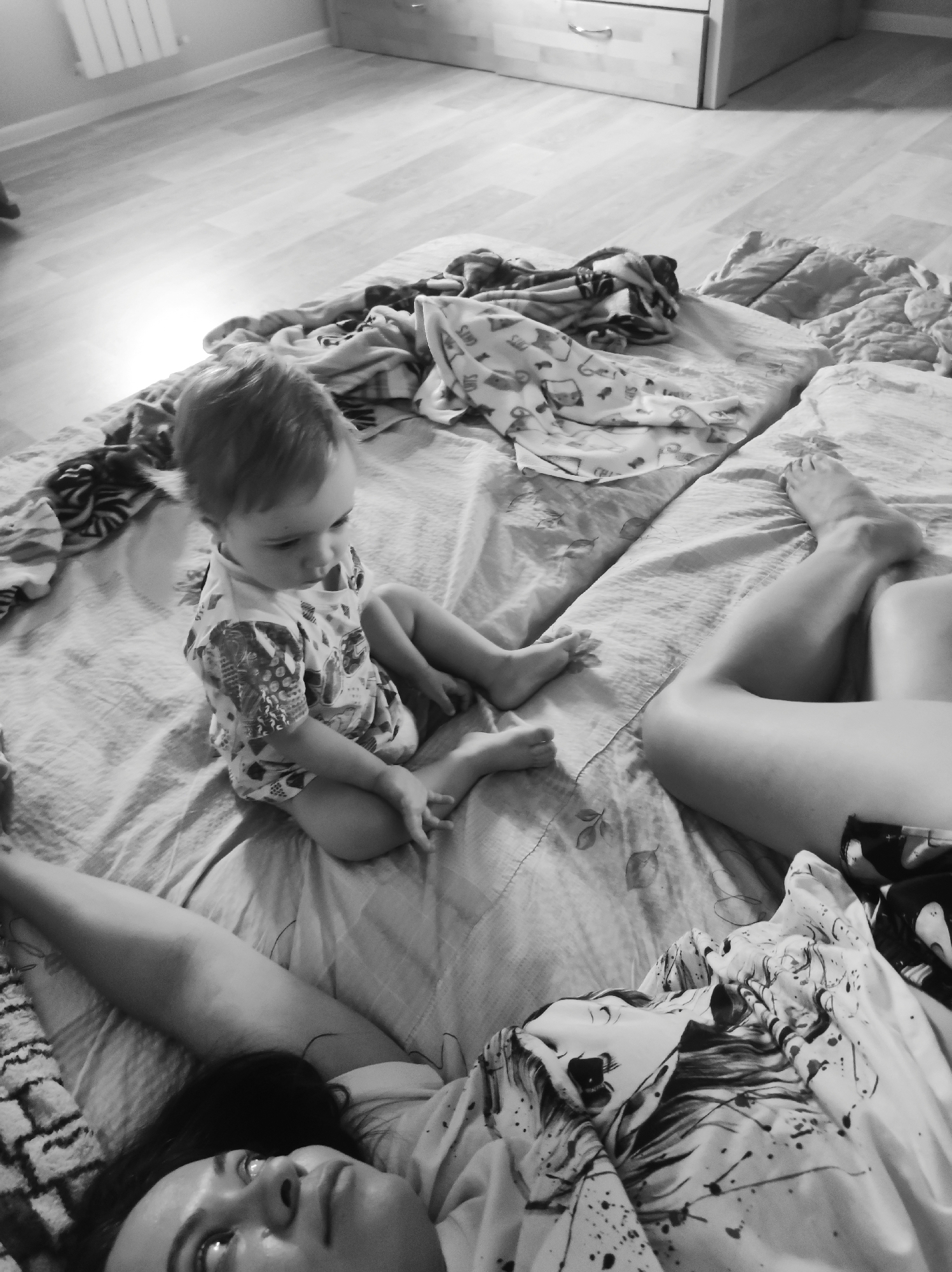 A black and white photograph depicts a baby sitting cross-legged on a bed with a woman lying down next to them. The baby is wearing a patterned onesie and is looking down. The woman's face is visible, and she appears to be looking upwards. The bed is covered in a patterned sheet and some clothing. The room also contains a radiator and a chest of drawers. The location is unknown.
