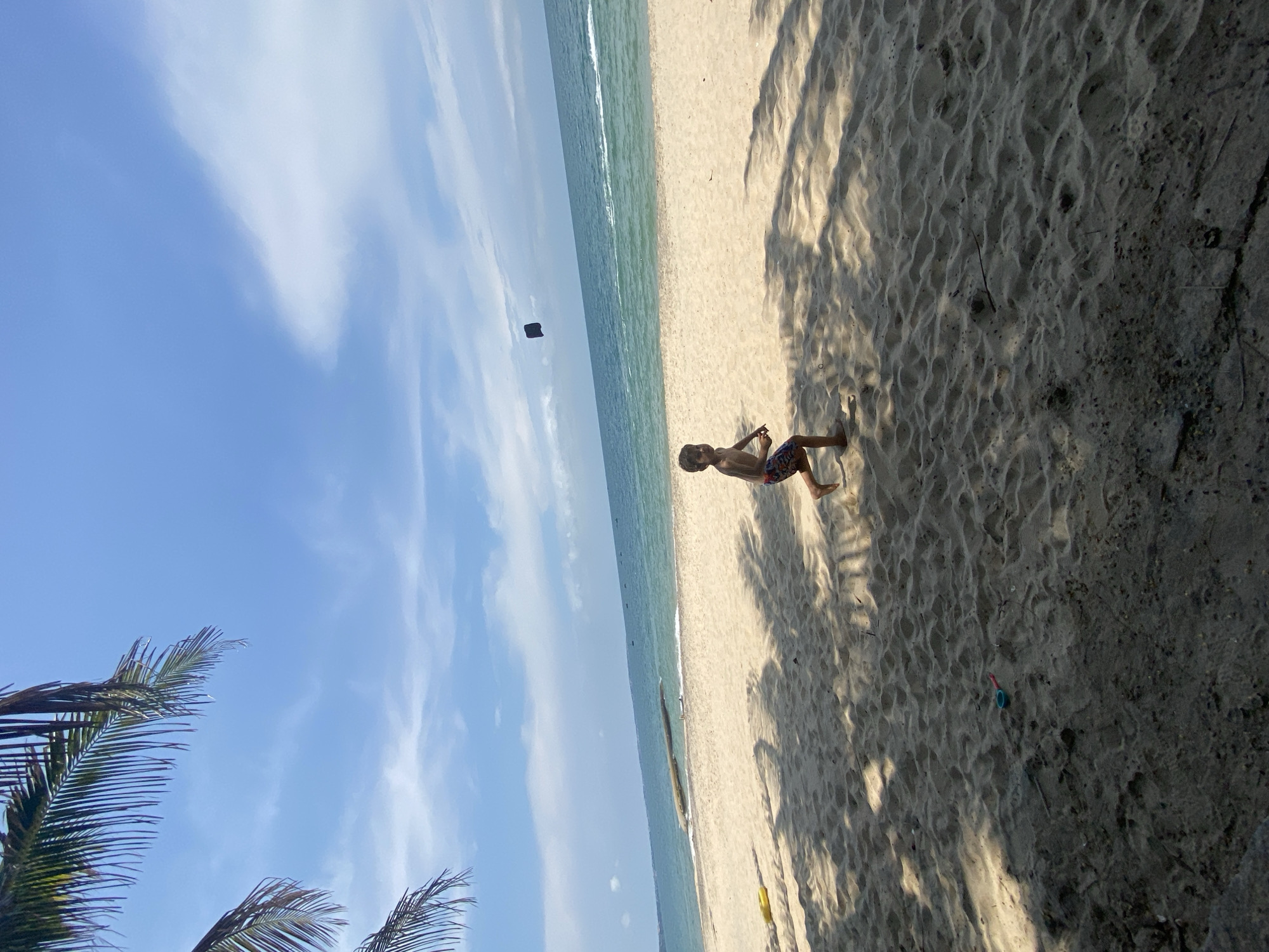 A young boy is running on a sandy beach with the ocean in the background. The boy is wearing patterned shorts. In the sky above the boy, there is a dark object. Palm fronds are visible in the upper left corner of the image, casting shadows on the sand. The beach is wide and appears to be relatively empty. The water is a calm blue, with some small waves breaking near the shore. The sky is blue with scattered white clouds. The scene is brightly lit by sunlight, suggesting it is daytime. The location is identified as Phu Thuy Ward, Vietnam, a coastal area known for its beaches.
