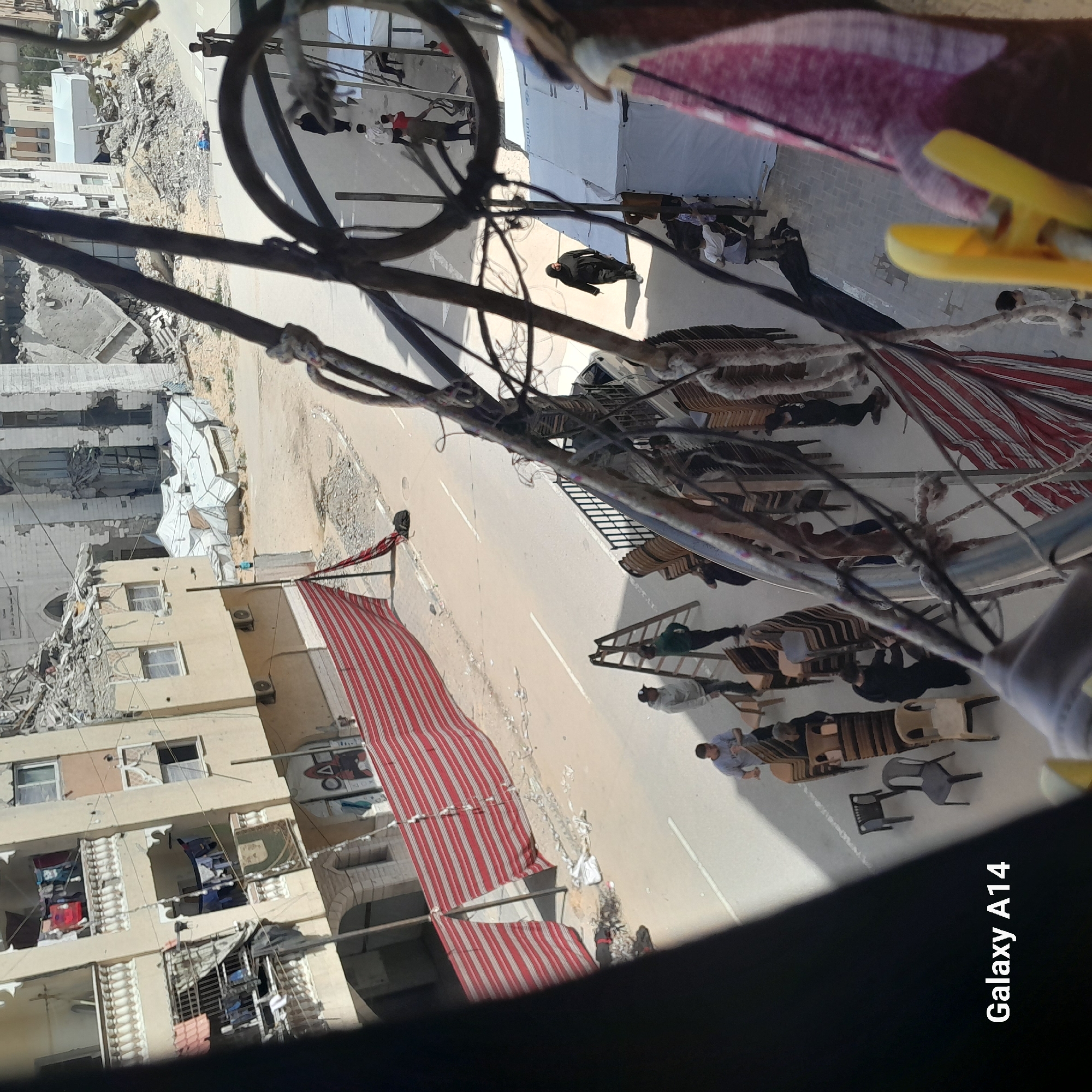 The image, oriented sideways, captures a bustling street scene from an elevated perspective, likely from a balcony or window, with dark cables and a yellow clothes peg prominent in the immediate foreground.
When viewed correctly, the street extends from the foreground into the background, flanked by buildings. On the left, several multi-story buildings appear significantly damaged, with visible rubble and collapsed sections indicating structural compromise. Adjacent to one of these damaged structures is a white tent with the word "UNICEF" clearly visible, suggesting a humanitarian presence. Further down, intact apartment balconies display clothes hanging to dry. A long, red and white striped awning extends over a section of the street from one of the buildings.
The street itself is sunlit, featuring a paved surface with white lines. Numerous individuals are present, engaged in various activities. Many people are gathered around, sitting on or standing near stacks of plastic and wooden chairs arranged along the thoroughfare. One person is visible ascending a wooden ladder, while another figure appears to be lying on the ground further up the street. A vehicle is partially seen in the background.
The strong shadows suggest bright daylight. The combination of damaged infrastructure and the humanitarian aid tent implies a setting affected by conflict or disaster. The bottom right corner of the image contains the text "Galaxy A14", indicating the camera used.
