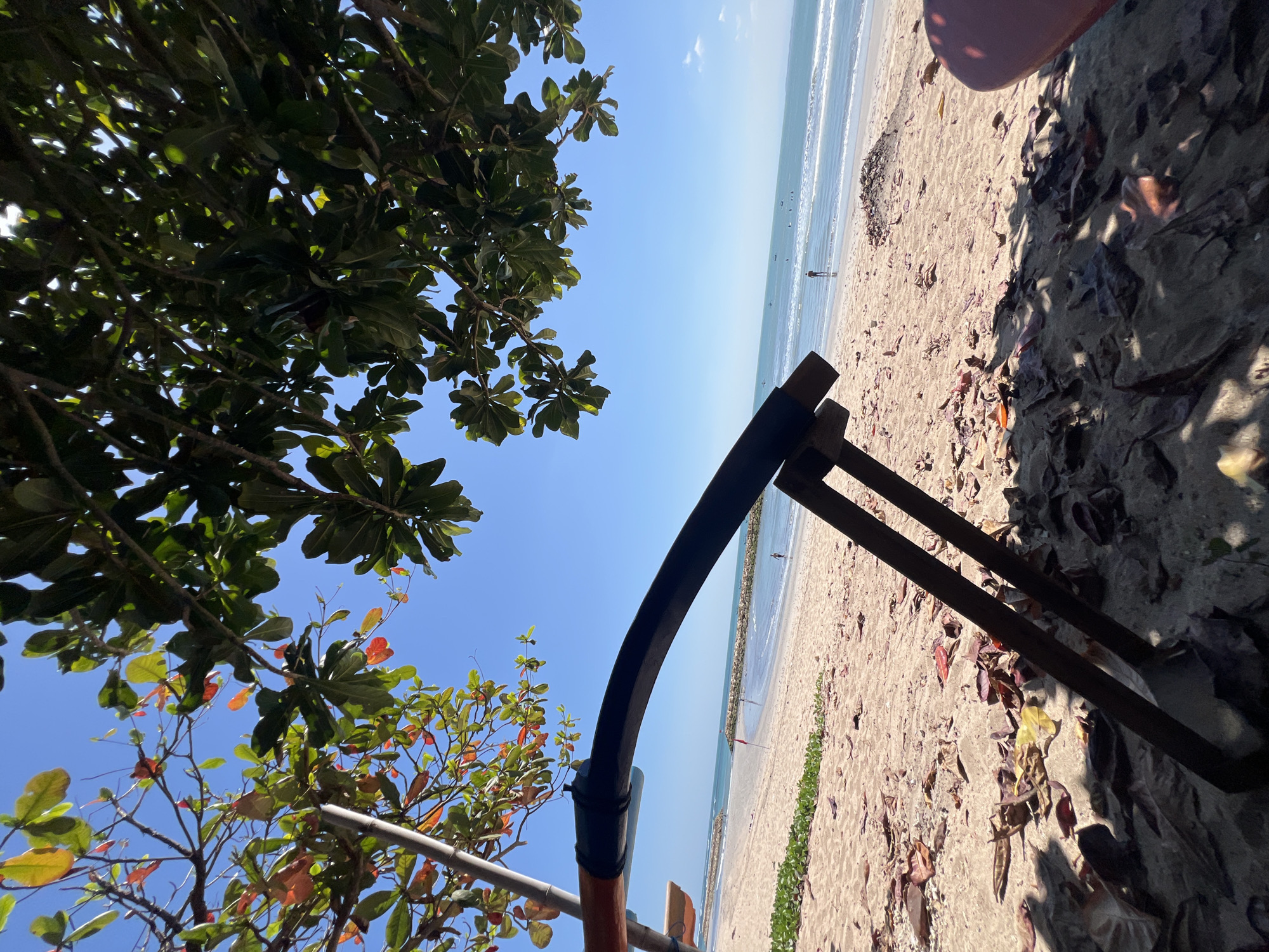 The image depicts a beach scene in Kuta, Indonesia, on a clear day with a bright blue sky. In the foreground, the focus is on the sandy beach with fallen leaves scattered across it. A part of a wooden beach chair or sun lounger is visible, casting a shadow. Lush green foliage from trees overhangs the scene from the top. In the mid-ground, the sandy beach stretches towards the ocean, with a few people walking or standing on the shore. A red flag on a pole is planted in the sand. Further out, the turquoise ocean meets the horizon, with gentle waves lapping the shore. Several small boats are visible on the water, and a lone figure is standing in the ocean, appearing to be wading. The overall impression is one of a serene and sunny day at a tropical beach.