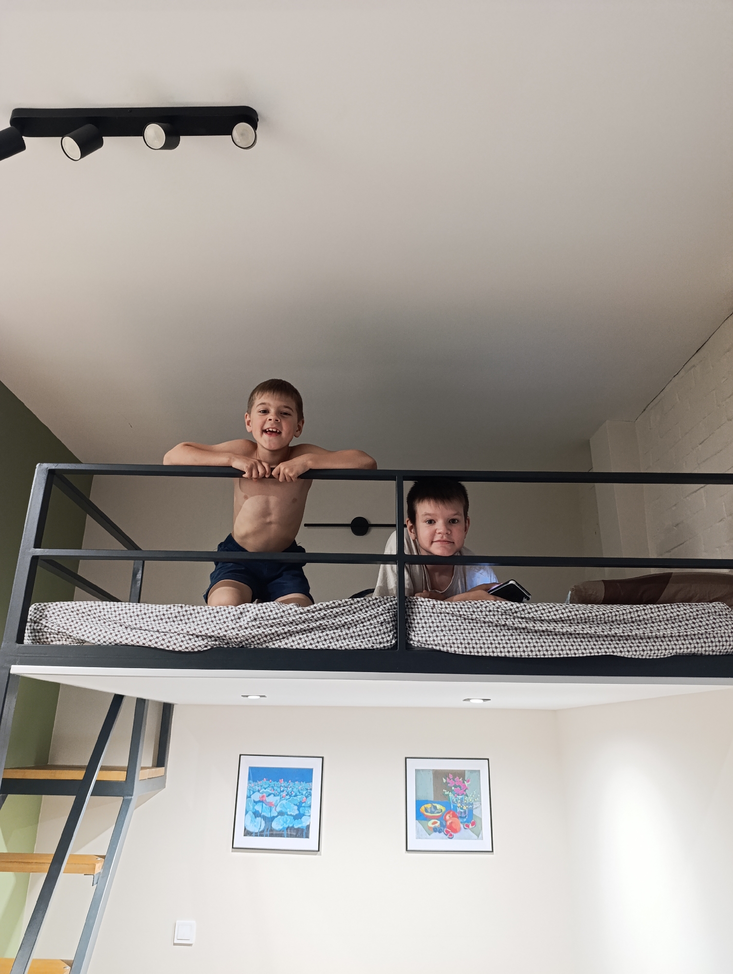 Two young boys are seen from below, looking down from a black metal-framed loft bed. The boy on the left, shirtless and wearing dark blue shorts, beams with a wide, joyful smile, leaning forward on the bed's railing. His arms rest on the top bar as he looks directly at the viewer. To his right, a second boy, wearing a light-colored shirt, also leans over the railing, holding a smartphone in his hands. He offers a more subdued smile, engaging with the camera.
The loft bed features a black and white patterned duvet or blanket, and a robust black metal frame with several horizontal bars forming the railing. To the left, a staircase with wooden steps and a black metal frame leads up to the bed.
The room's ceiling is white, featuring a modern black track light fixture with multiple cylindrical spotlights. Below the loft, the walls are predominantly cream or off-white. Two framed artworks hang on the wall: the left one depicts blue water lilies, and the right one is a still life with fruit and flowers in a vase. A small white light switch and a recessed ceiling light are also visible near the paintings. Part of the wall to the left is painted an olive-green color, while a section on the right shows a subtle white brick-like texture. Behind the second boy, a black, circular wall-mounted fixture or decoration is visible.
The scene conveys a playful and happy atmosphere, likely indoors during daytime hours, with bright, even lighting. There is no visible text in the image.