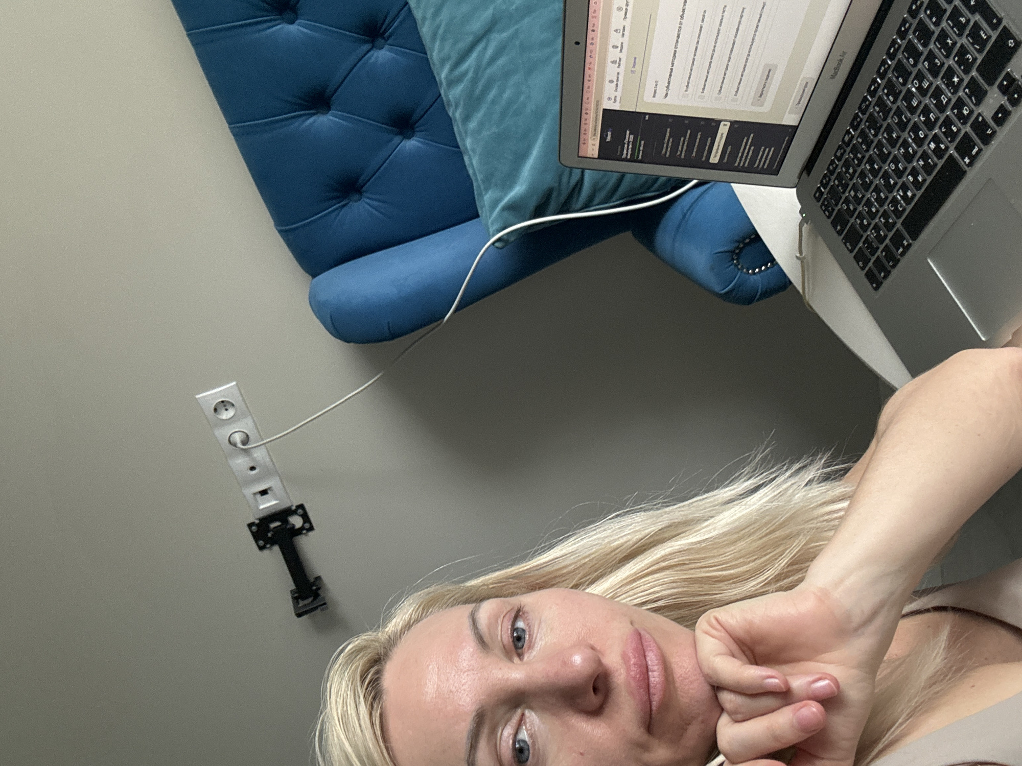 The image is a selfie taken by a fair-skinned woman with blonde hair, blue eyes, and pink lips. She is wearing a beige top. Her left hand is near her chin, and the focus is on her face.
In the background, a blue tufted armchair is visible. A laptop computer, presumably a Macbook Air, is resting on the chair, with its screen displaying content. A white cable is connected to the laptop.
On the wall, there's a power outlet with multiple sockets, and a black bracket is attached to the wall below the outlet. The background wall is painted a light gray color. The location is Unknown, Unknown.