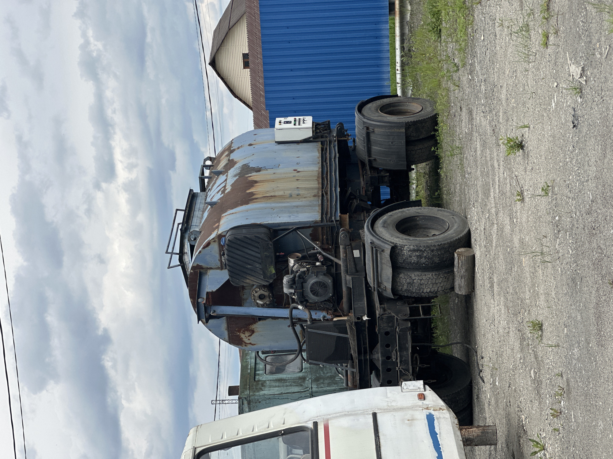An old, rusty blue tanker truck is parked on a gravel lot next to a white vehicle, likely a bus. The tanker truck has a large cylindrical tank, a visible engine, and multiple tires. In the background, a blue corrugated metal fence and a house with a brown roof are visible. The sky is overcast with gray clouds. This scene is located in Slyudyanka, Russia.