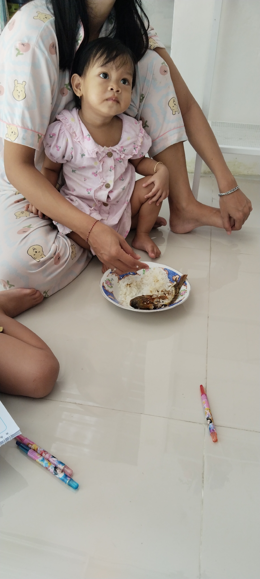 The image captures a scene from a home environment, likely in Kecamatan Pati, Indonesia. A young child is seated on the lap of an adult, both wearing matching pajama sets featuring cartoon bear and fruit prints. The child, wearing a pink ruffled dress with a cherry print, looks to the right with a curious expression. In front of them, on the floor, is a plate of white rice with a piece of cooked fish. To the right of the plate, a woman's bare feet are visible, one adorned with a bracelet. On the floor in the foreground, several pens with cartoon prints are scattered, along with an open notebook. The flooring appears to be light-colored tiles. The overall impression is one of a casual, domestic moment.