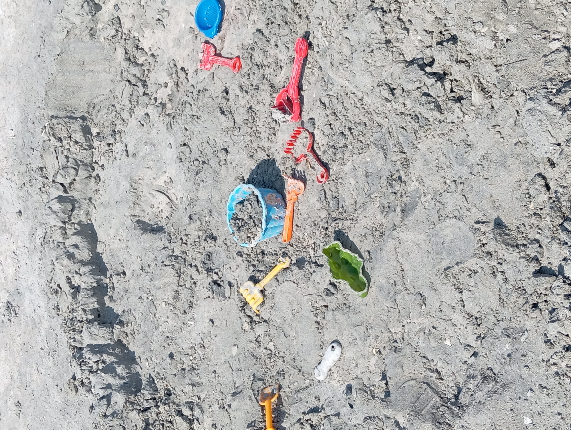 This is a ground-level, eye-level shot of a sandy area, likely a beach or sandbox, with several children's toys scattered across the surface. The sand is damp and compacted in some areas, with footprints visible. The toys include a blue plastic bucket filled with sand, a green plastic dinosaur-shaped mold, a yellow plastic rake and shovel, and several red plastic rakes and shovels. The lighting suggests it is daytime, possibly sunny. There are no people visible in the image. The scene is not located in Ömirzaq, Kazakhstan, as the provided image does not contain any discernible landmarks or context to suggest this location.