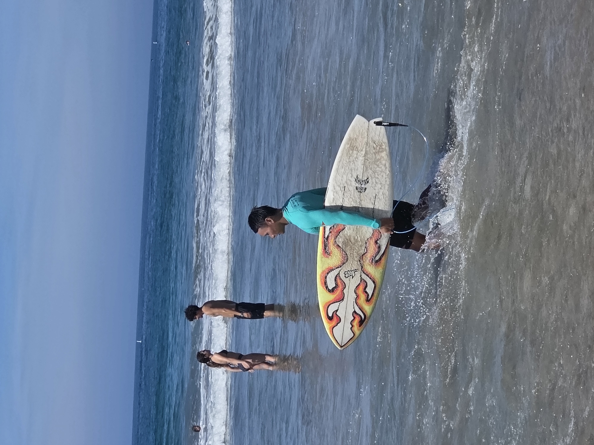 A man in a teal long-sleeved rash guard and black board shorts carries a surfboard with a flame design into the ocean at Santa Monica State Beach in California. The surfboard is white with red, orange, and yellow flames. The man is walking through shallow, choppy water, kicking up spray. In the background, a man and a woman stand in the water, facing each other. The woman wears a black bikini, and the man wears black swim trunks. The ocean is blue with white-capped waves. The sky is clear and blue, with a few distant boats visible on the horizon. The beach is not visible in this frame.