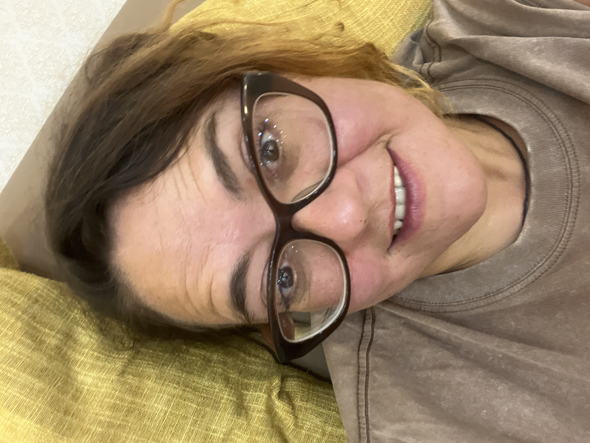 A woman with brown hair, wearing glasses and a brown t-shirt, is lying on a yellow textured surface, possibly a couch or pillows. She is looking directly at the camera with a slight smile. The image is a close-up, focusing on her face and upper torso. The background consists of the yellow material and a hint of a wall behind her. There is no information available about her location in Nha Trang Ward, Vietnam.