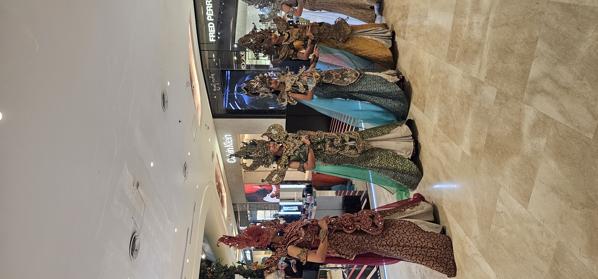 In a brightly lit, modern indoor location with a tiled floor in Denpasar, Indonesia, a line of four traditionally costumed female dancers, possibly performers or participants in a cultural event, are lined up horizontally. The dancers are adorned in ornate, colorful garments and headdresses featuring detailed patterns and embellishments. Behind them are several store fronts including Fred Perry and Calvin Klein. The background suggests a shopping mall or similar commercial space.