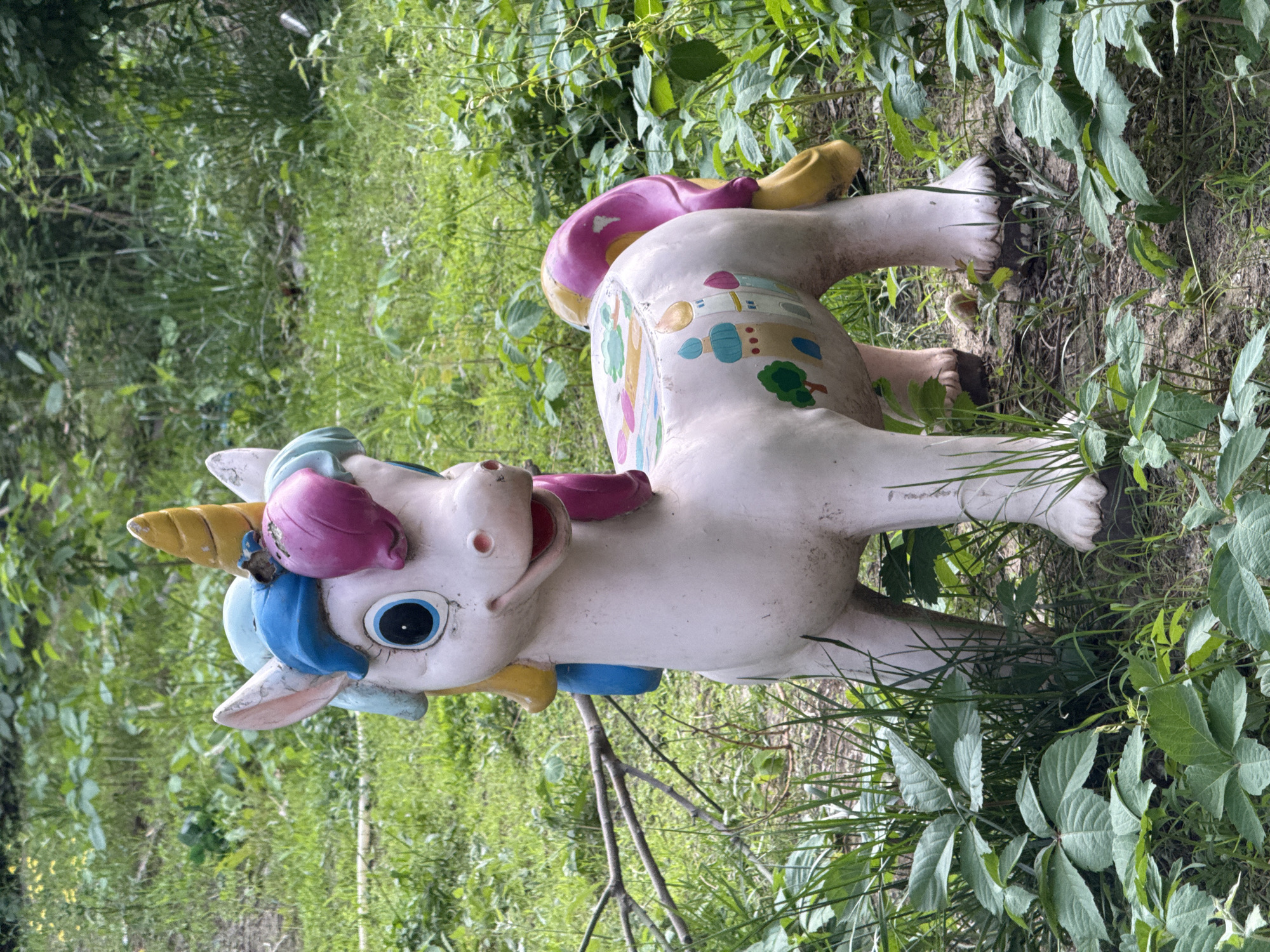 A weathered, faded white unicorn statue stands amidst dense green foliage. The statue is painted with colorful details on its body, including a pink tail, blue and yellow mane, and painted images of buildings and trees on its torso. The statue's paint is chipped and aged. It is positioned in a garden or overgrown area in Chaoyang District, China.
