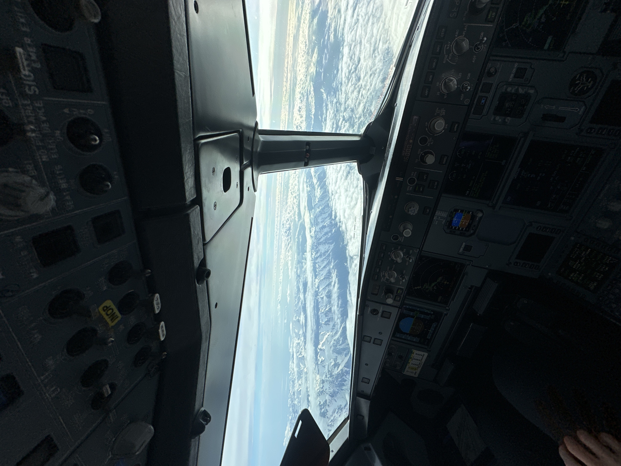 The image is an interior shot from the cockpit of an Airbus aircraft in flight, looking out over a landscape with mountains and clouds. The control panels and instrumentation of the cockpit are visible on both sides of the frame. The landscape visible through the windshield appears to be mountainous, with areas of snow cover and cloud cover. It's not possible to determine the location as Sochi, Russia from the information provided in the image. There are no people visible.