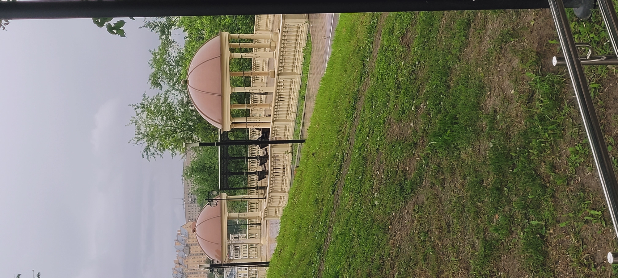 The image depicts a park setting in Unknown, Unknown, featuring two domed structures resembling pavilions. The pavilions are constructed with light-colored columns and a decorative base, topped with copper-colored domes. They appear to be part of a larger architectural design, possibly a temple complex or public monument. The park's environment is characterized by green grass, suggesting a well-maintained landscape. The sky is overcast, casting a soft, diffused light over the scene. An indistinct urban landscape is visible in the background, indicating the park's location within the city. Metal railings are seen in the foreground, possibly part of a walkway or barrier within the park. No people are visible in the immediate scene.