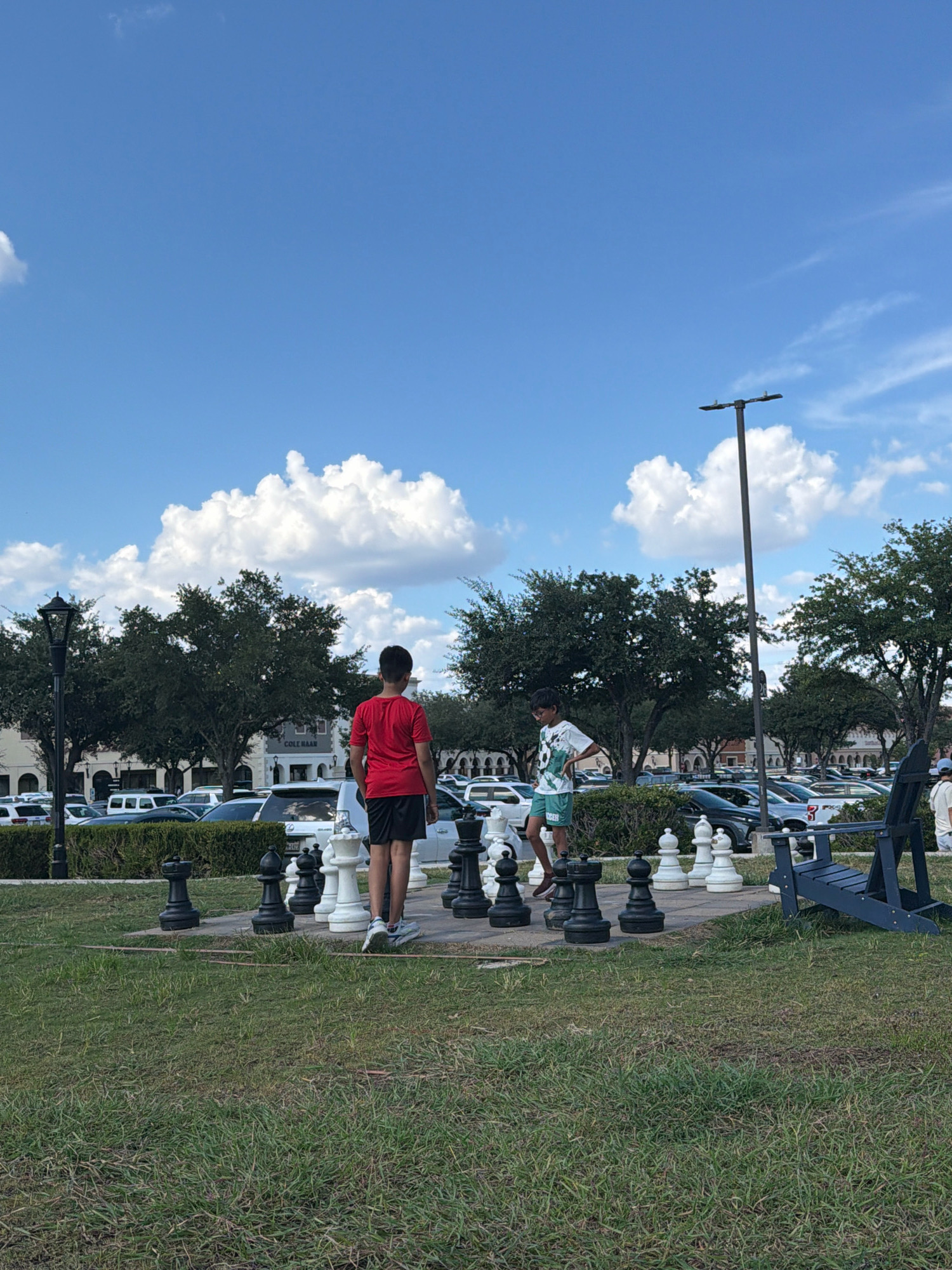 In San Marcos, Texas, USA, two young individuals are engaged in a game of giant chess outdoors. The scene is set on a grassy area adjacent to a parking lot filled with vehicles and a row of buildings. One person, wearing a red t-shirt and black shorts, stands to the left, facing away from the camera and towards the chessboard. The other, in a patterned t-shirt and green shorts, is positioned to the right, looking at the chessboard. The giant chess pieces are prominently featured on a paved surface, with a blue Adirondack chair visible to the right of the chessboard. Trees line the background, and a lamppost stands behind the chess players. The sky above is blue with scattered white clouds.